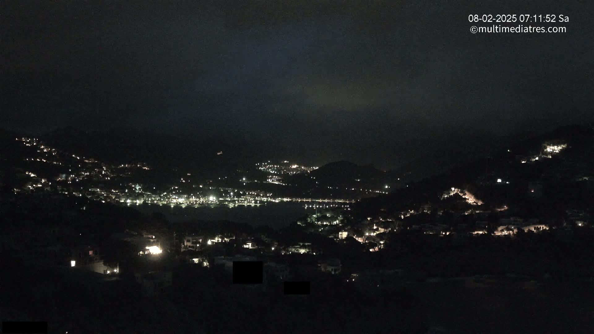 A nighttime view of a coastal town nestled in hills, with scattered lights illuminating the landscape under a dark, cloudy sky.
