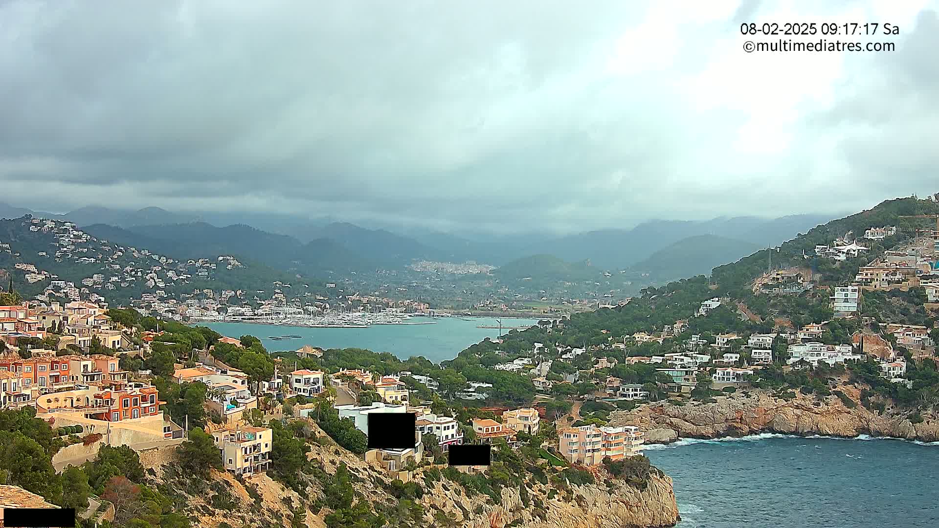 A coastal town with numerous houses clings to a hillside overlooking a calm bay under an overcast sky.