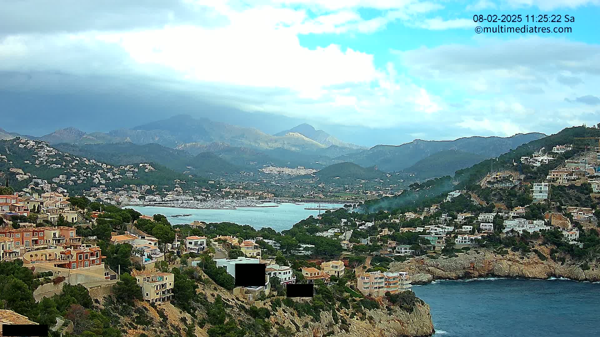 A coastal town nestled in a mountainous region under a partly cloudy sky overlooks a calm bay filled with boats.