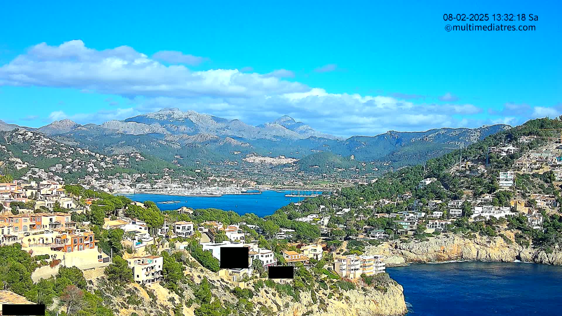 A coastal town nestled on a hillside overlooking a calm bay, with mountains in the background under a partly cloudy sky.