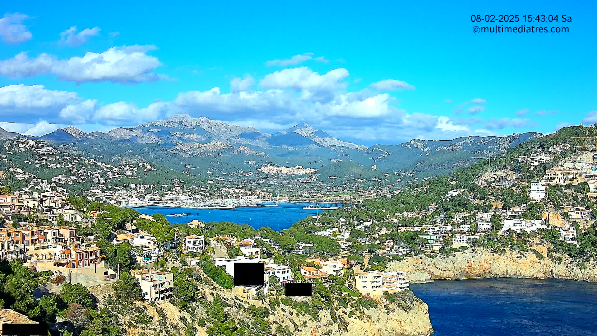 A coastal town nestled amongst hills and mountains under a partly cloudy blue sky overlooks a calm bay with many boats.