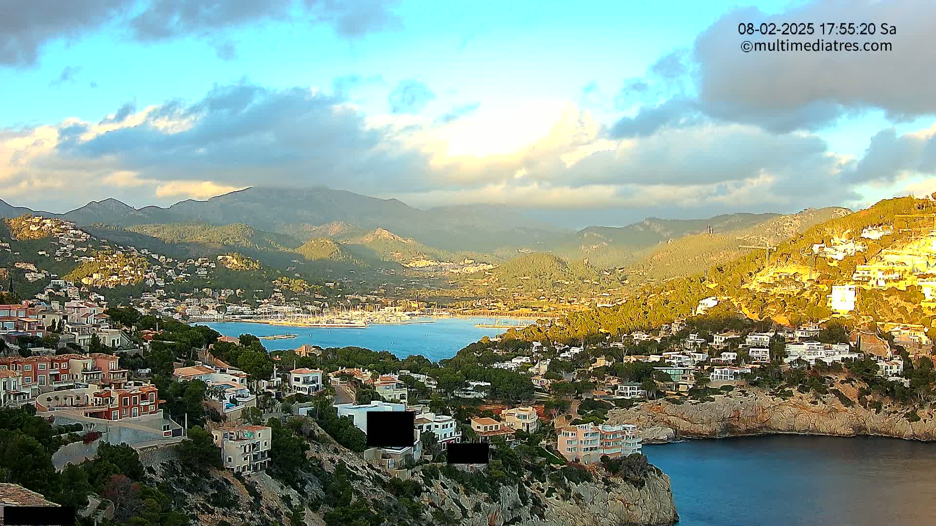 A coastal town nestled in a mountainous region, with a calm bay filled with boats, under a partly cloudy sky.