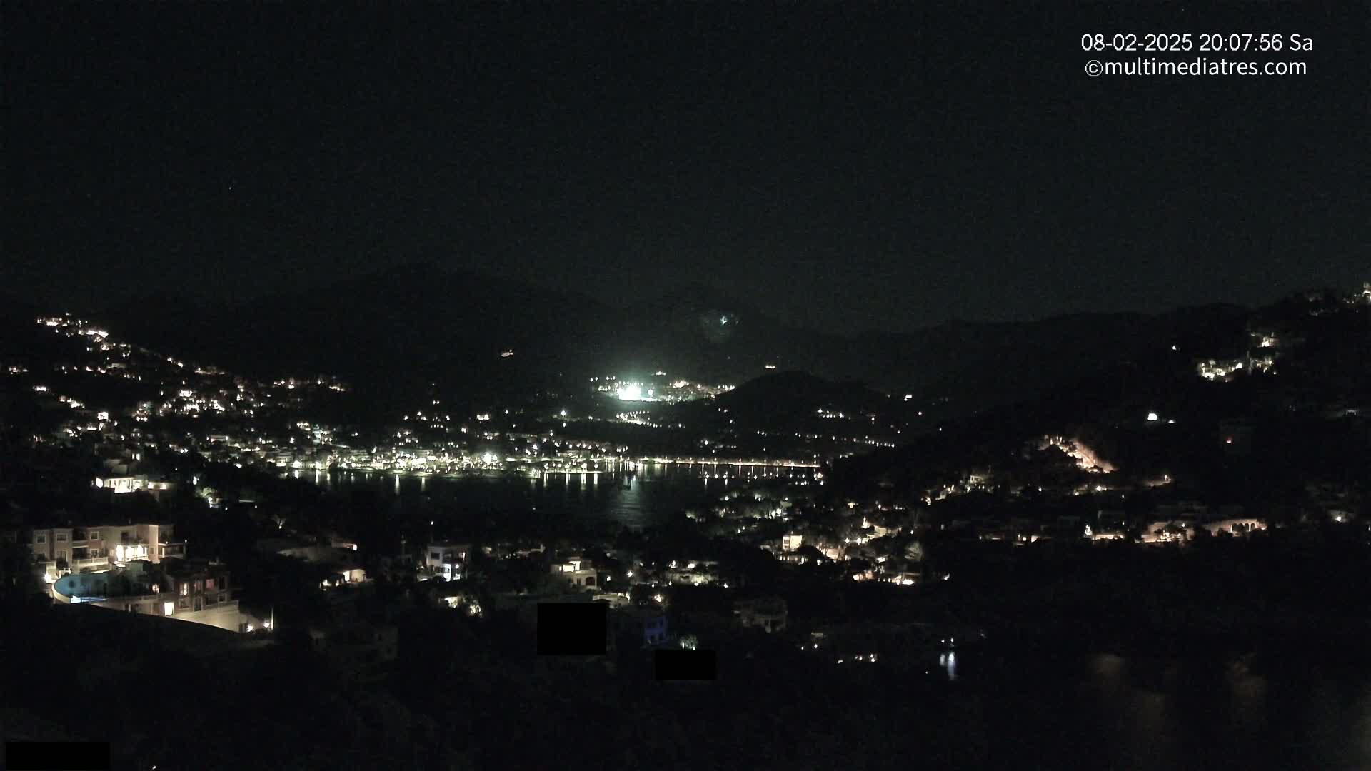 A nighttime view of a coastal town nestled between hills, with numerous lights illuminating buildings and a dark body of water separating the hillsides.