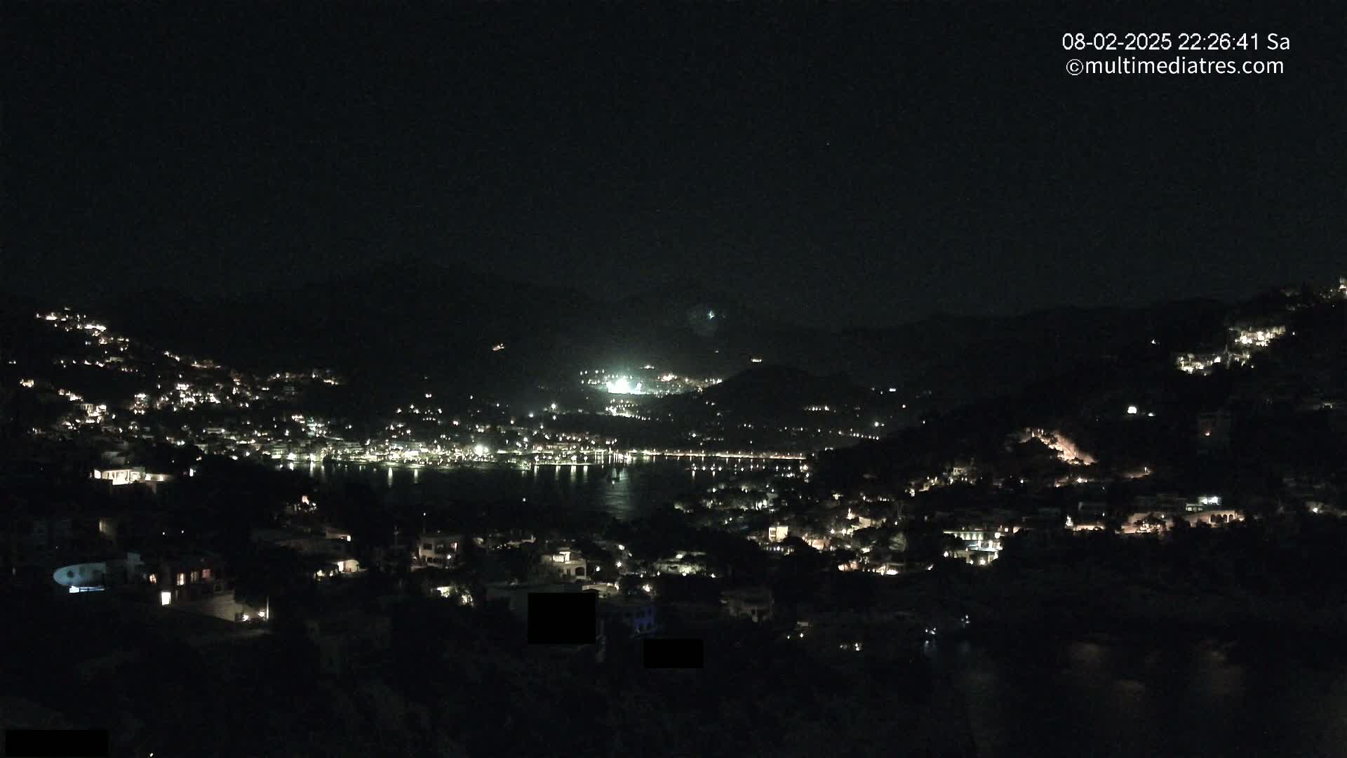 A nighttime view of a coastal town nestled between hills, with numerous lights illuminating the buildings and a dark body of water visible in the distance under a clear sky.