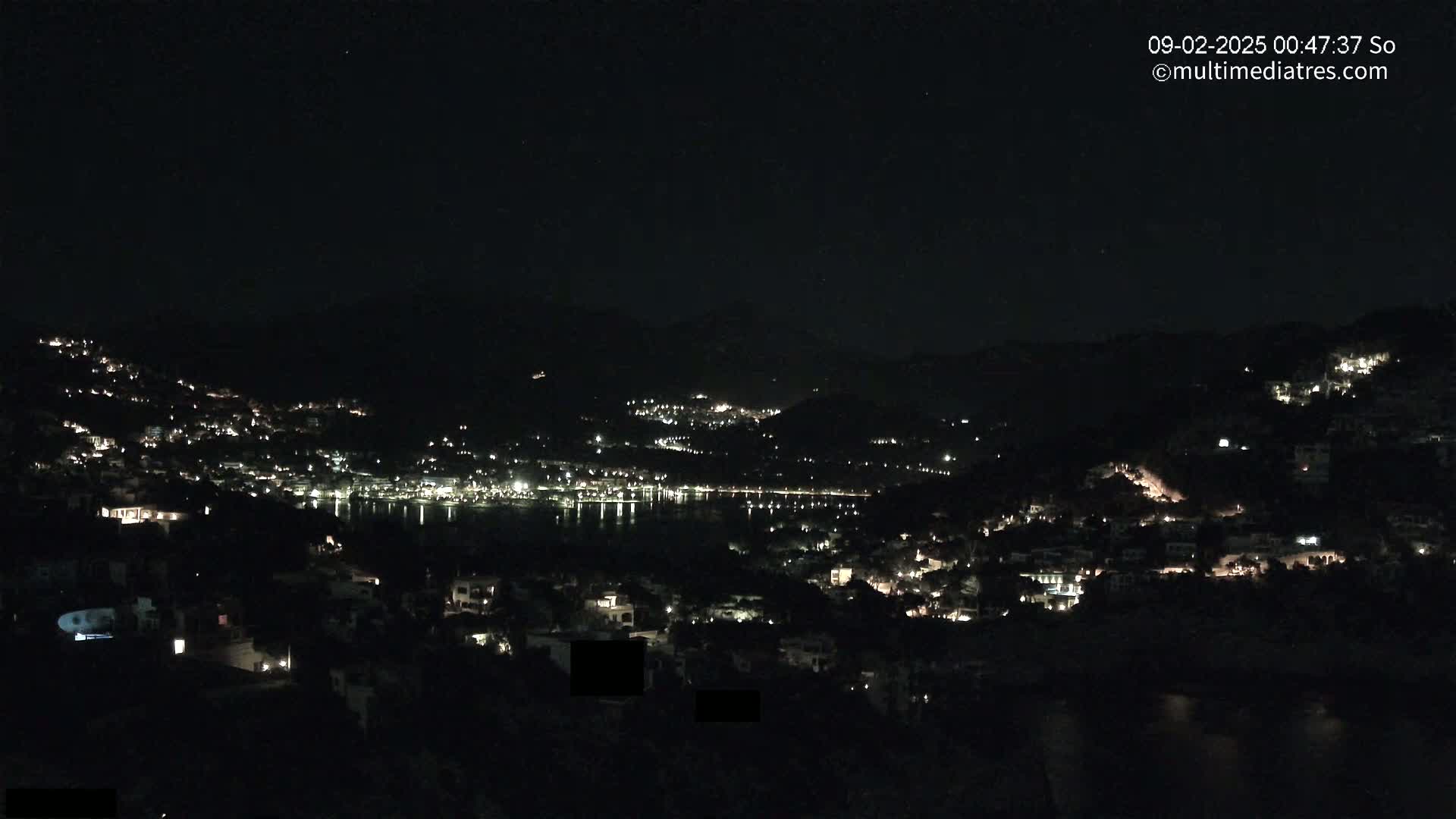 A nighttime view of a coastal town nestled on hillsides, with scattered lights illuminating the buildings and a dark body of water reflecting the lights.