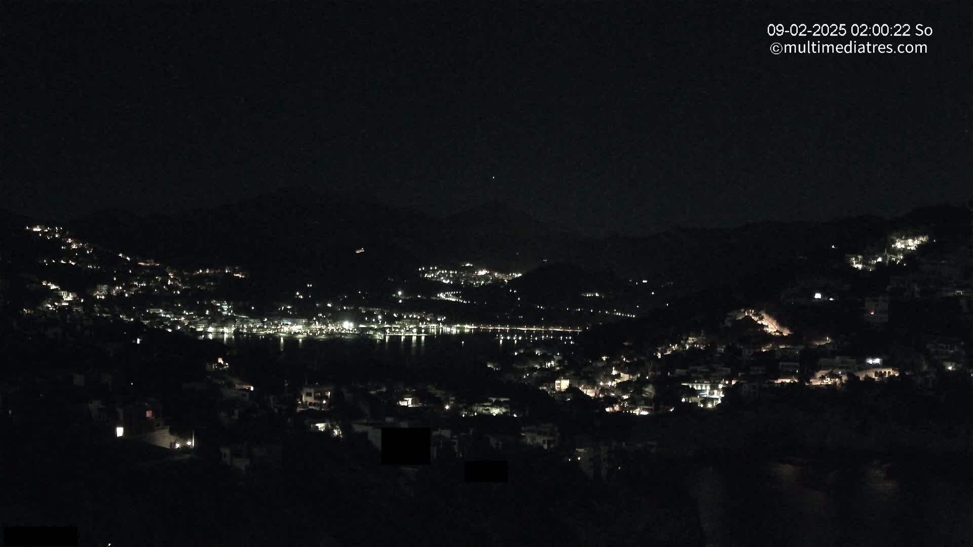 A nighttime view of a coastal town nestled among dark hills, with scattered lights illuminating the buildings and a calm body of water visible in the distance under a clear, dark sky.