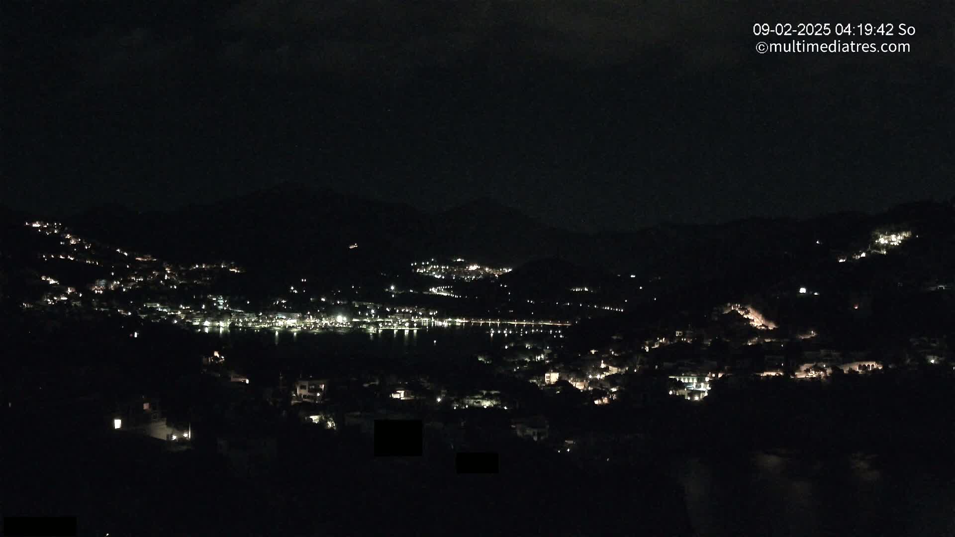 A nighttime view shows a coastal town nestled in dark hills, its lights twinkling along the shoreline and climbing the slopes.