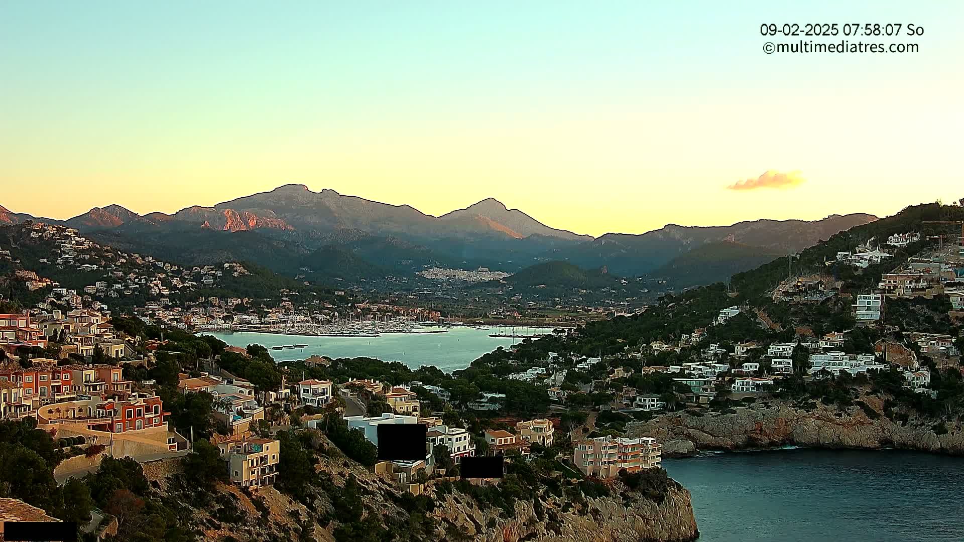 A coastal town nestled in a hillside overlooking a calm bay, with mountains in the background under a clear, sunrise sky.
