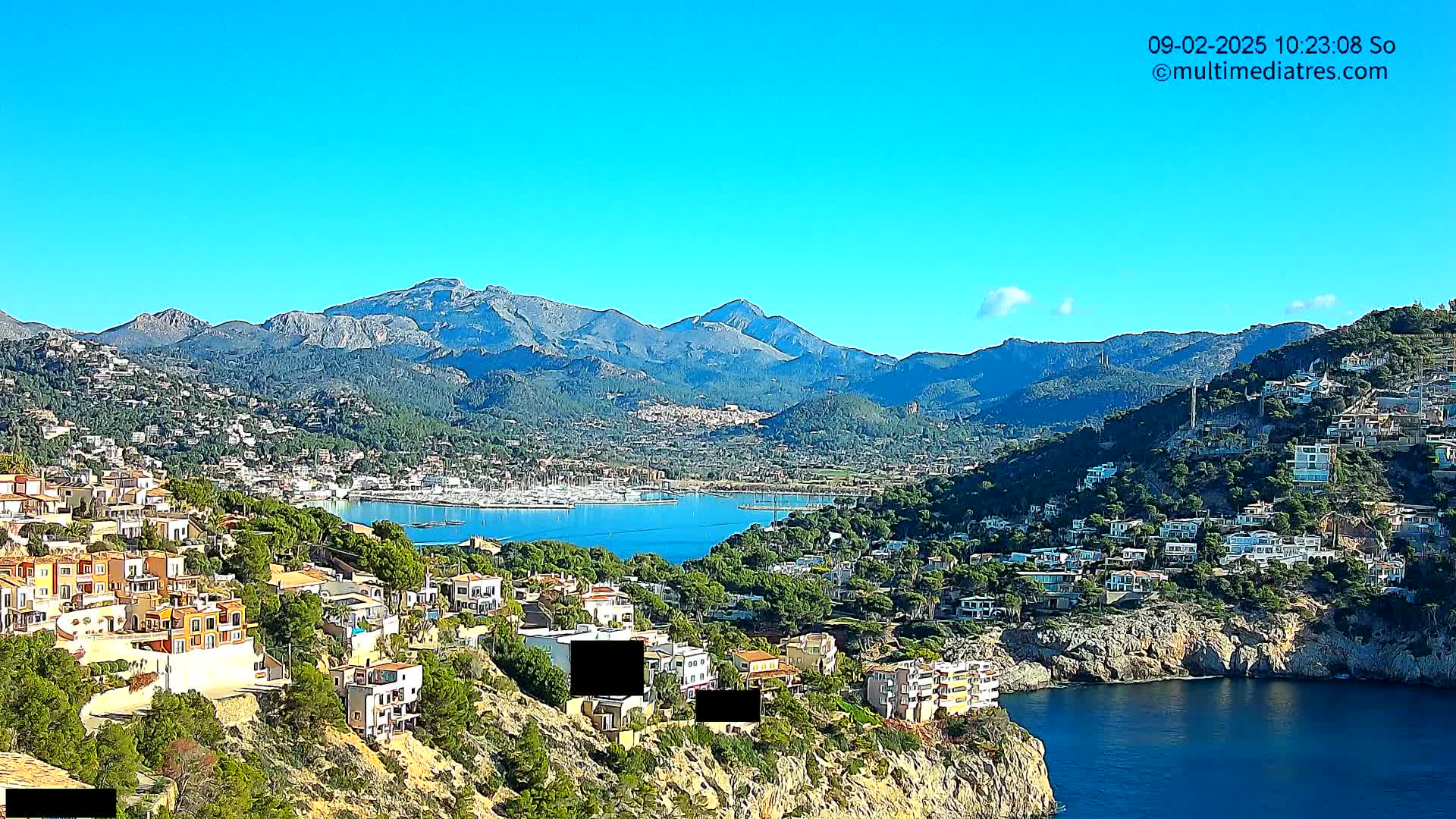 A coastal town nestled on a hillside overlooking a calm bay and a mountain range under a clear blue sky.