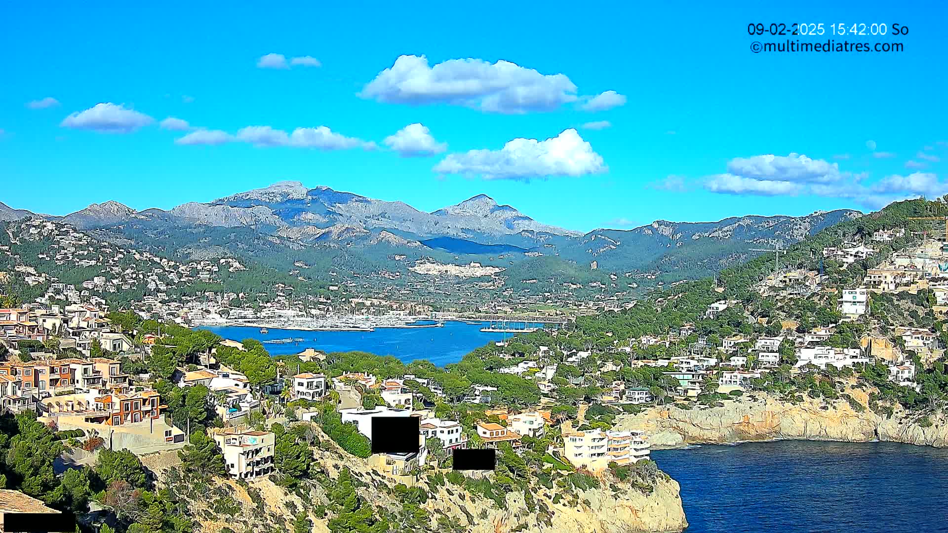 A coastal town nestled on a hillside overlooking a calm bay with numerous boats, all backed by a range of mountains under a mostly sunny sky with a few scattered clouds.