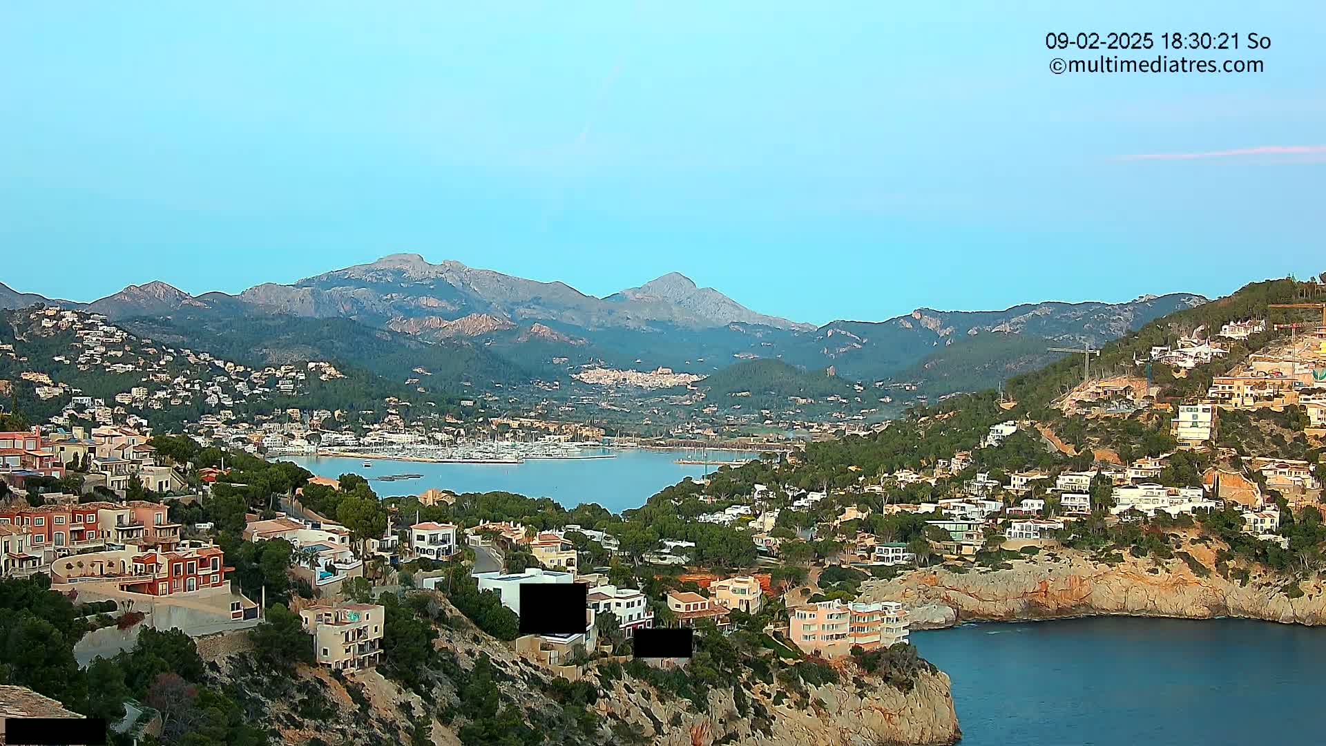 A coastal town nestled in a mountainous region, with a calm bay filled with boats, under a clear, light-blue sky.