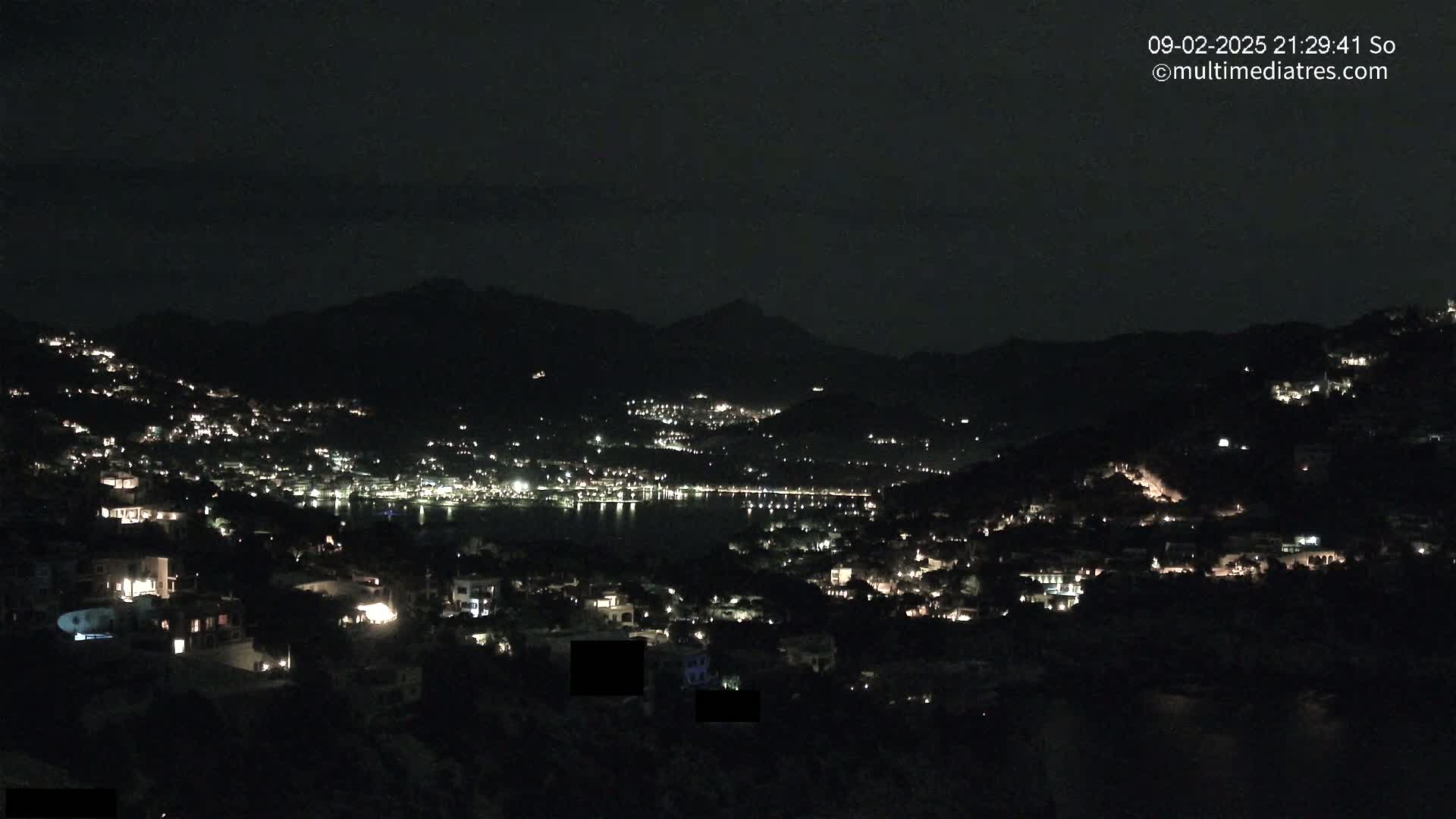 A nighttime view of a coastal town nestled in dark hills, with scattered lights illuminating buildings and a body of water reflecting the lights.