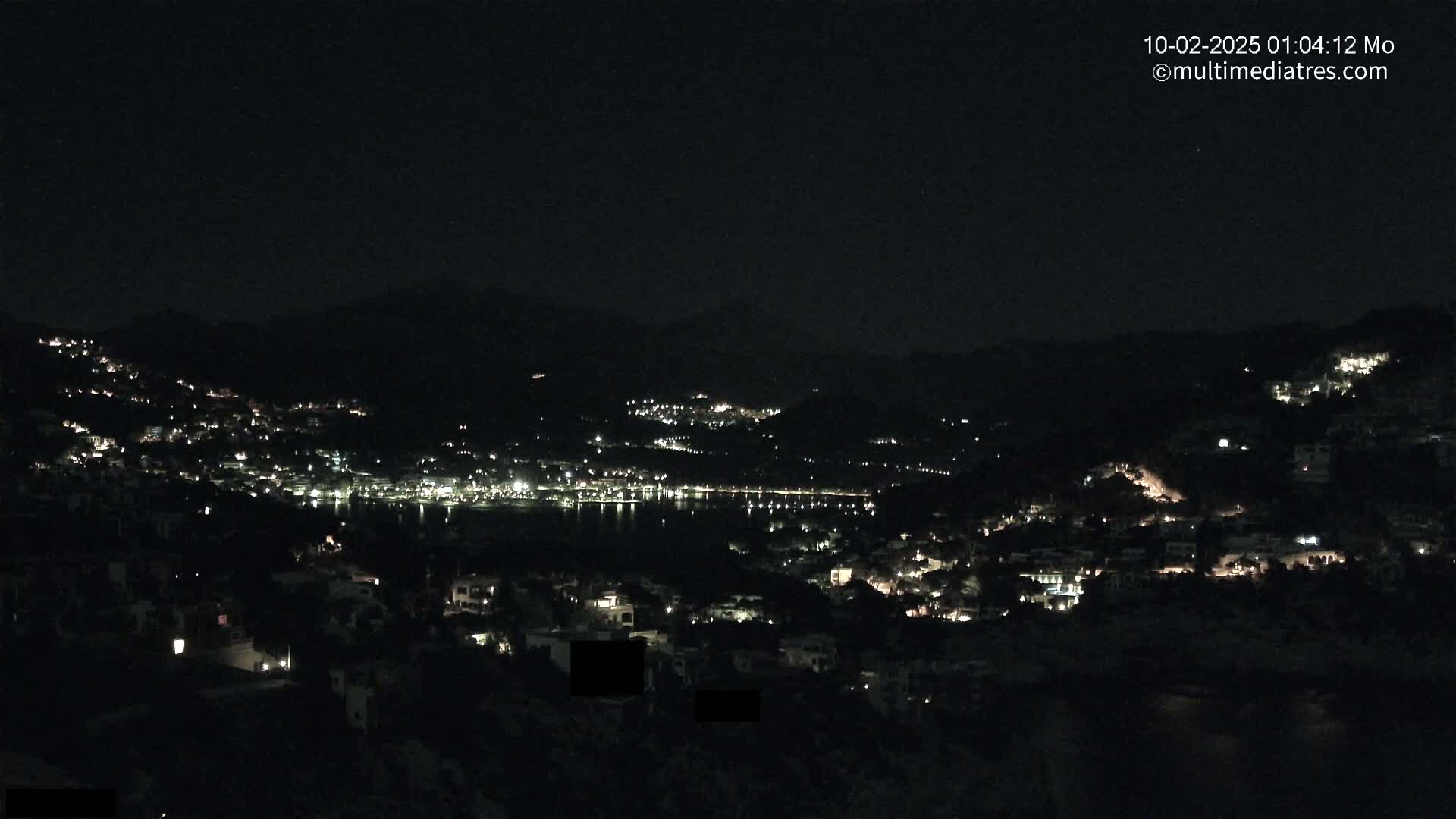 A nighttime view of a coastal town nestled on a hillside, with numerous lights illuminating buildings and a dark body of water visible in the distance under a clear sky.