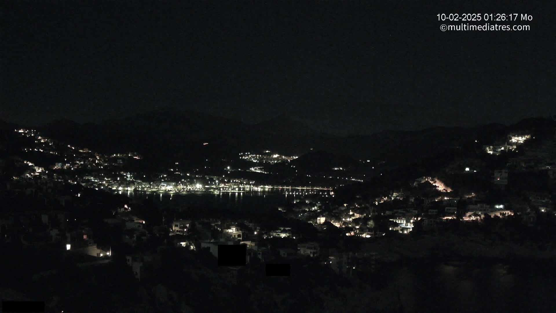 A nighttime view of a coastal town nestled in hills, with scattered lights illuminating the buildings and a dark body of water visible in the distance under a clear, dark sky.