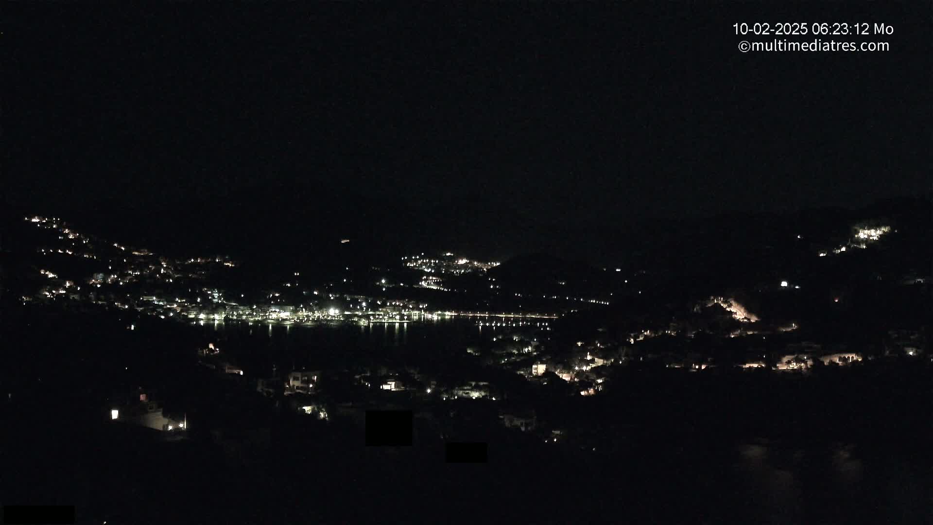 A nighttime, long-range view of a coastal town nestled among hills, with scattered lights illuminating the landscape.