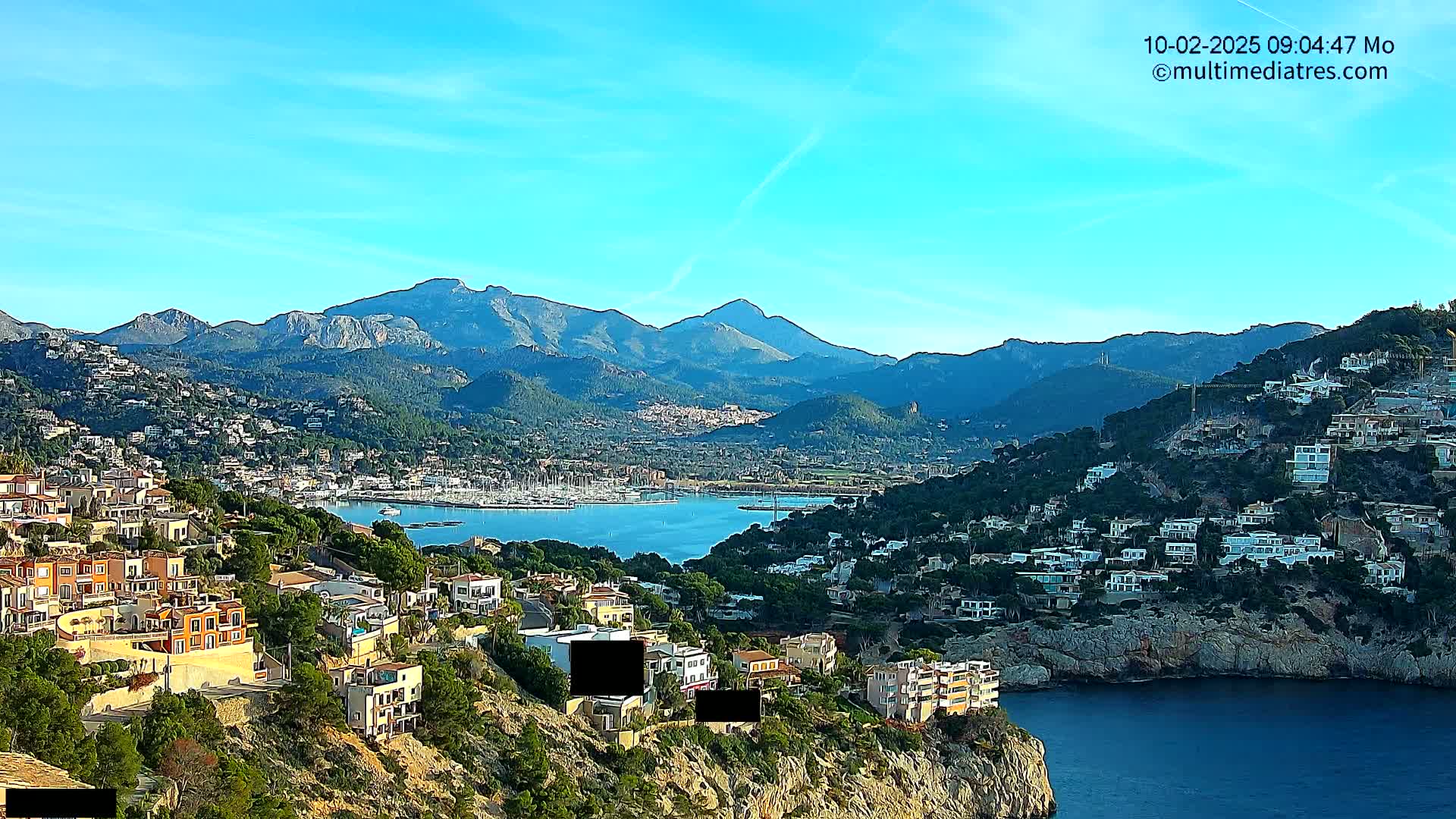 A coastal town nestled in a mountainous region under a clear blue sky, with a calm bay filled with boats visible in the distance.