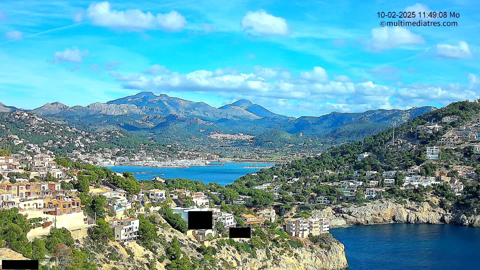 A coastal town nestled against a mountainous backdrop under a mostly sunny sky with scattered clouds.