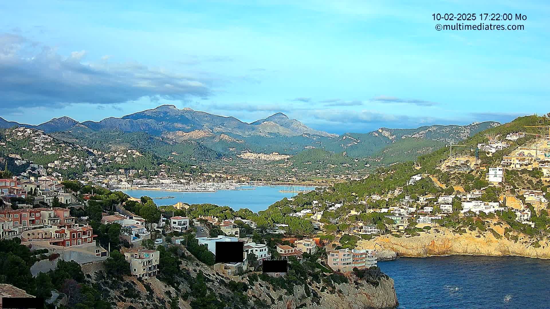 A coastal town nestled against a mountain range under a partly cloudy sky overlooks a calm bay filled with boats.