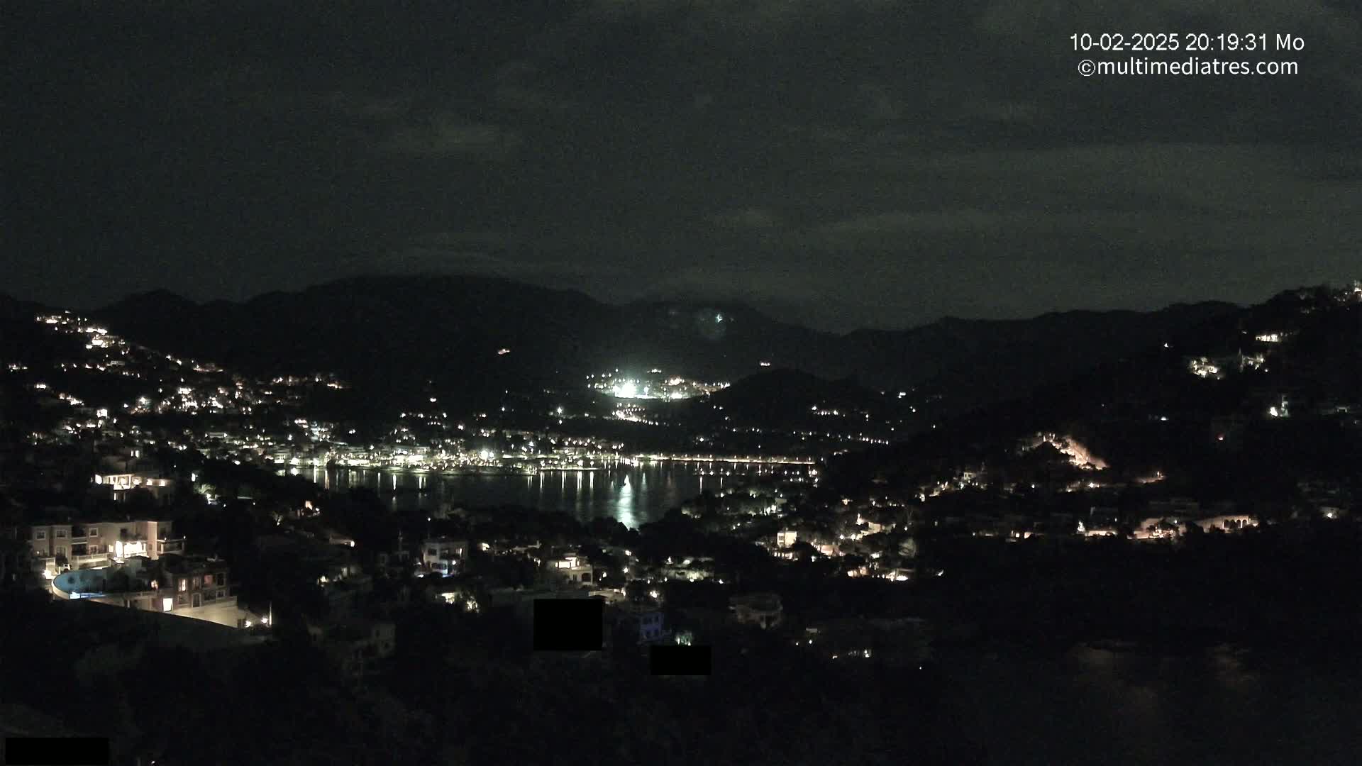 A nighttime view of a coastal town nestled between hills and a calm body of water, illuminated by scattered lights.