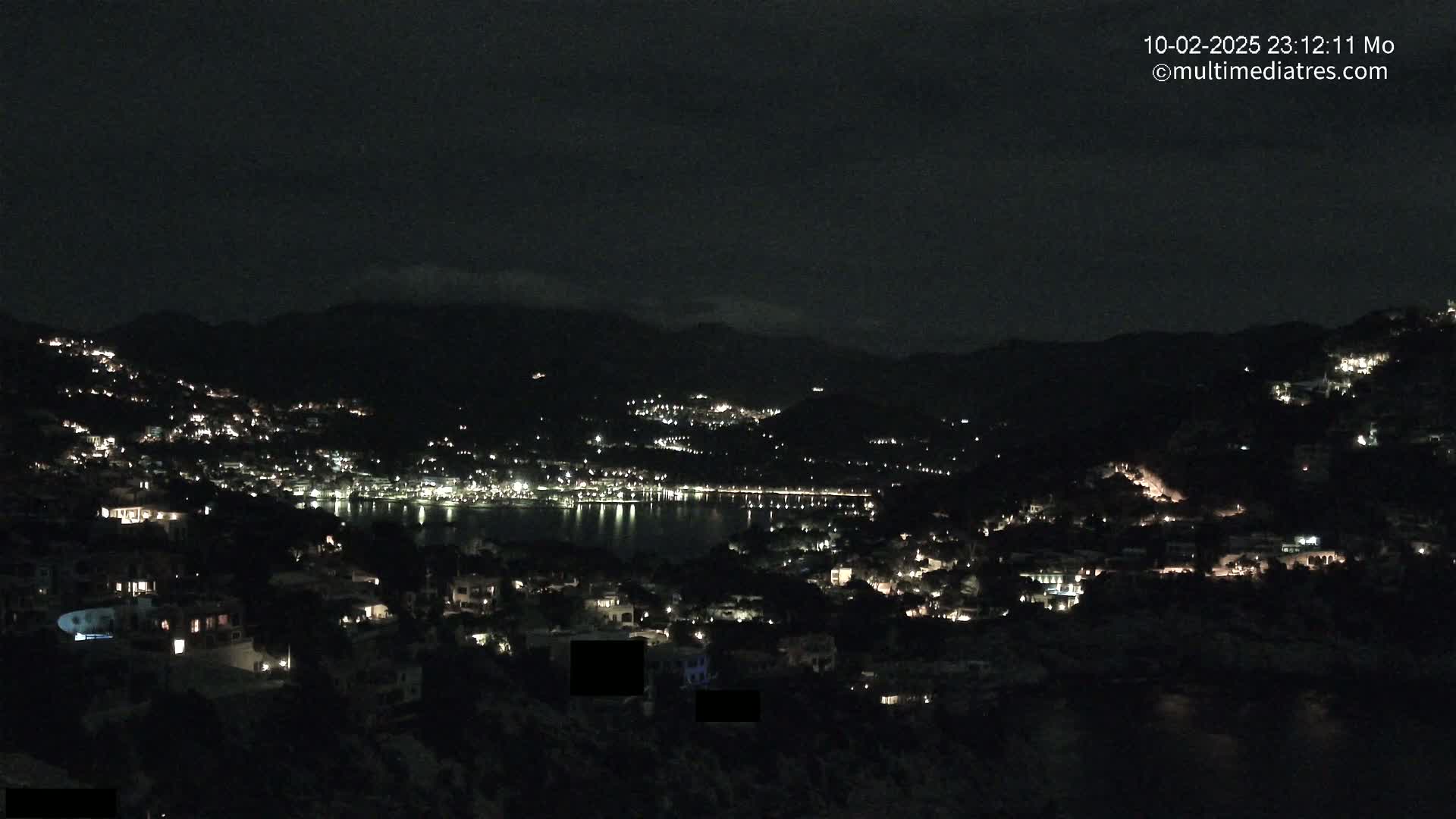 A nighttime view of a coastal town nestled in hills, with scattered lights illuminating the buildings and a dark body of water reflecting the lights.