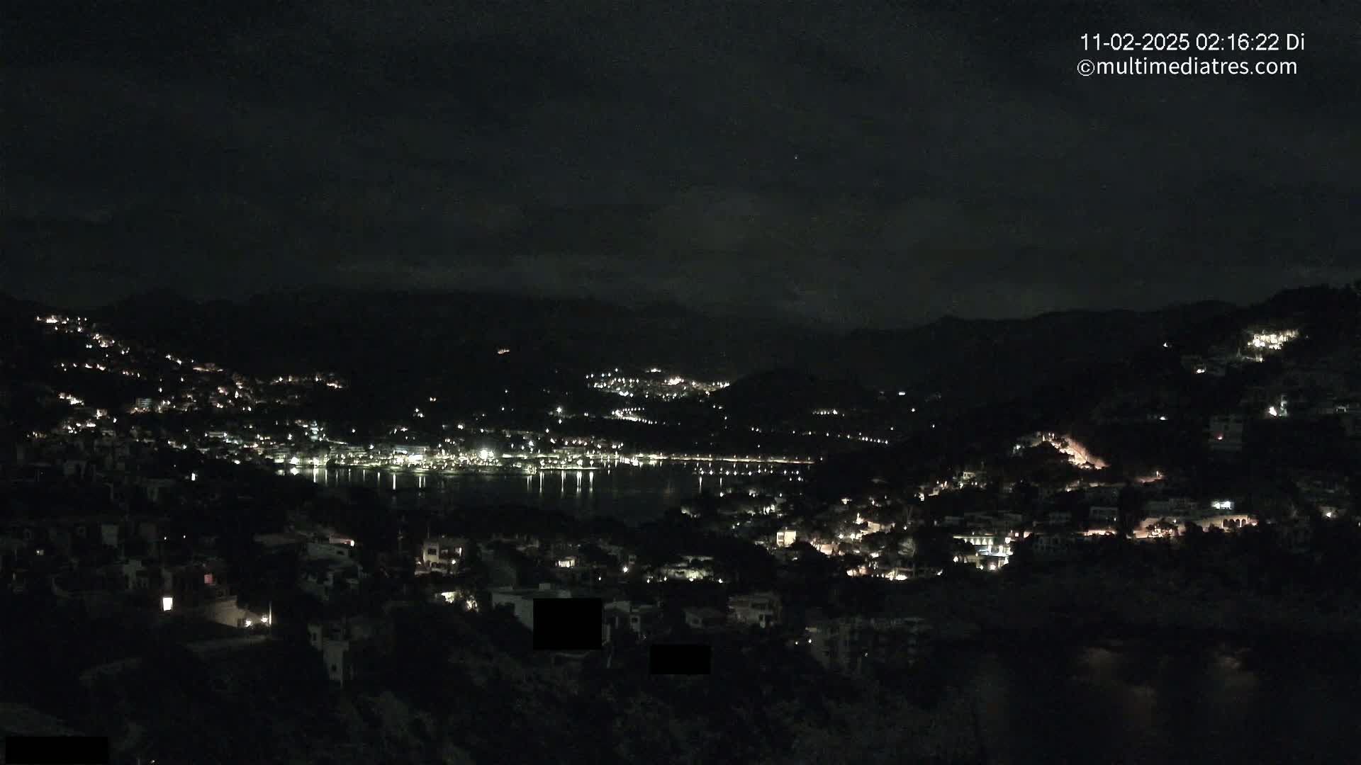A nighttime view of a coastal town nestled in hills, with scattered lights illuminating the landscape and a calm body of water reflecting the lights.