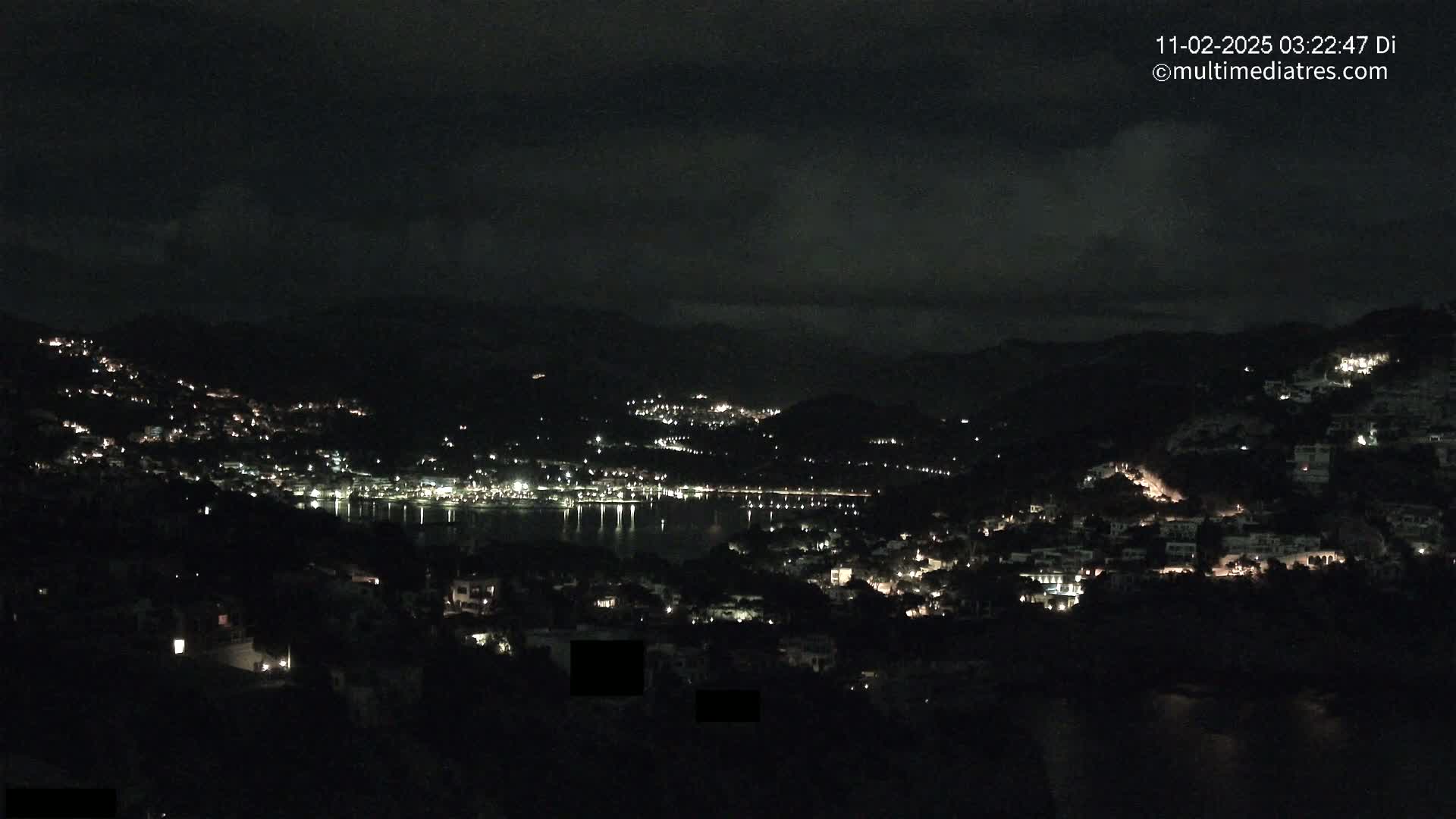 A nighttime view of a coastal town nestled between hills, with scattered lights illuminating the buildings and a calm body of water reflecting the lights.