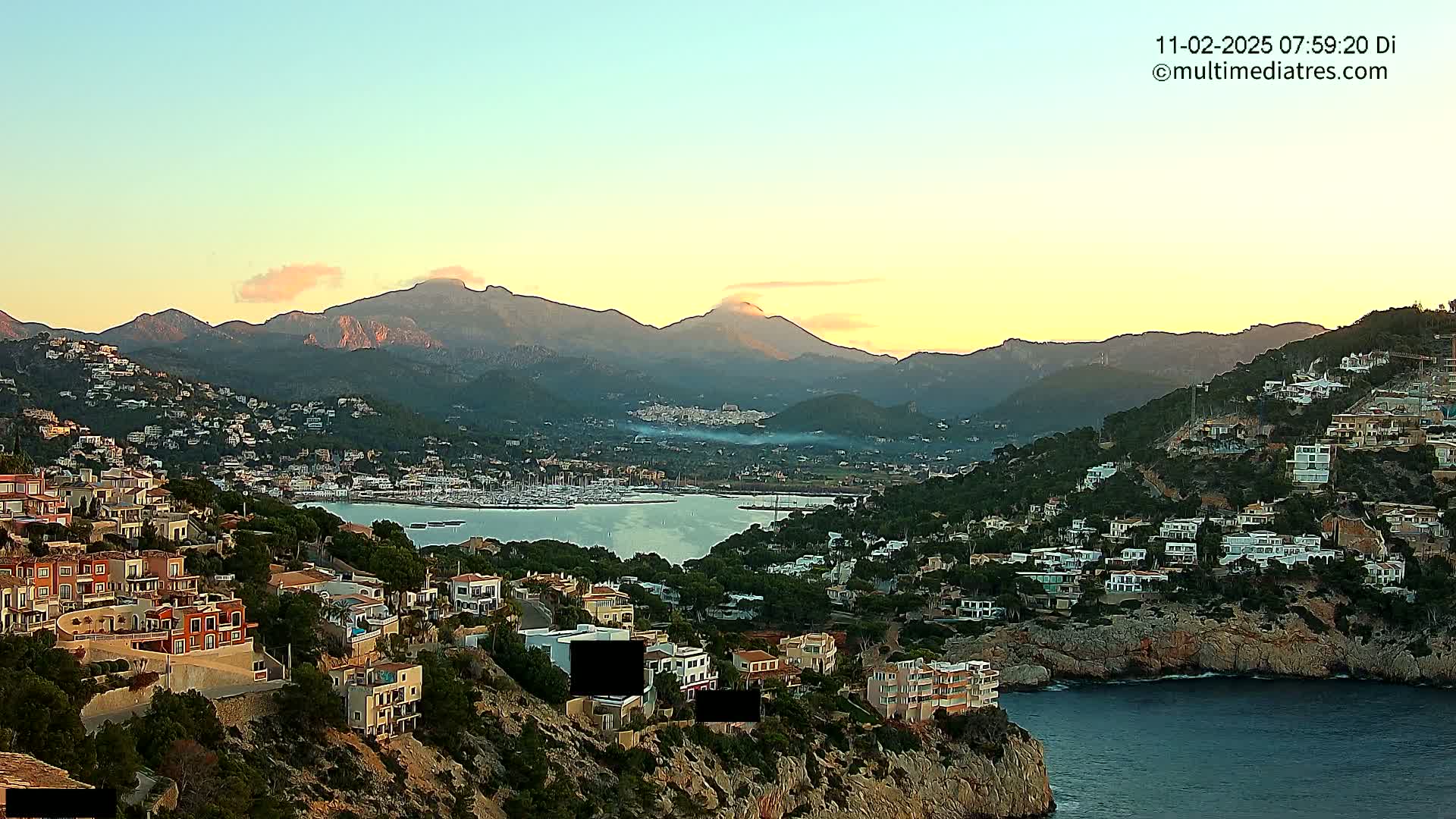 A coastal town nestled on a hillside at sunset, overlooking a calm bay filled with boats and backed by a mountain range under a clear sky.