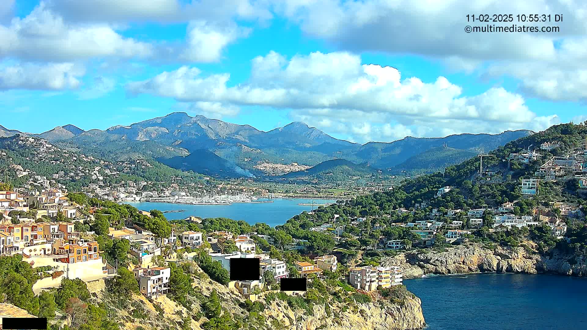 A coastal town nestled on a hillside, overlooking a calm bay with a marina and mountains in the background under a partly cloudy sky.
