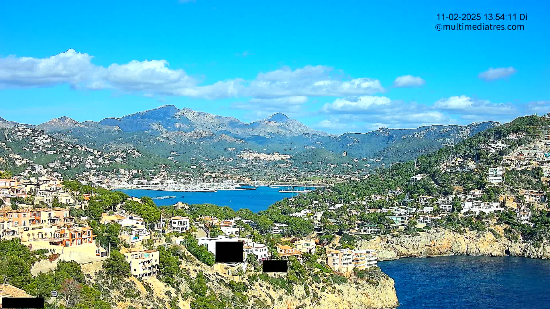 A coastal town nestled on a hillside overlooking a calm bay filled with boats, backed by a mountain range under a mostly sunny sky with scattered clouds.
