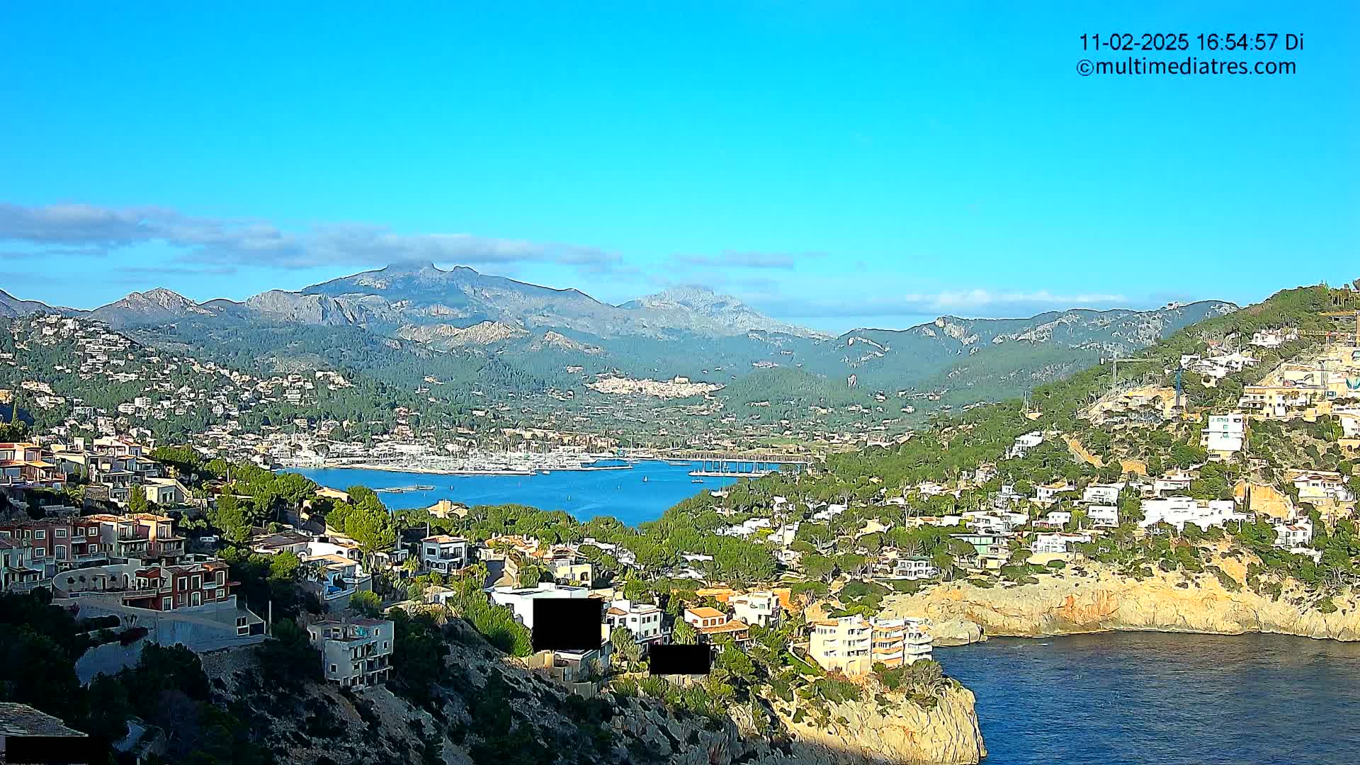 A coastal town nestled in a mountainous region overlooks a calm bay under a clear blue sky.