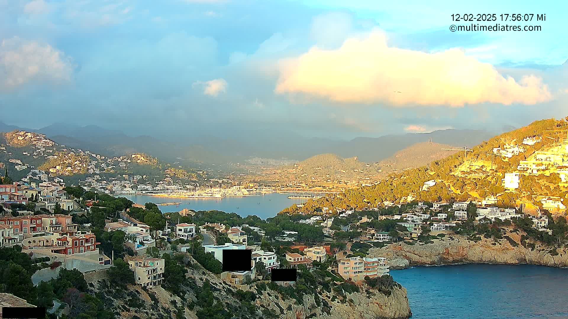 A coastal town nestled in hills overlooking a calm bay filled with boats, under a partly cloudy sky.