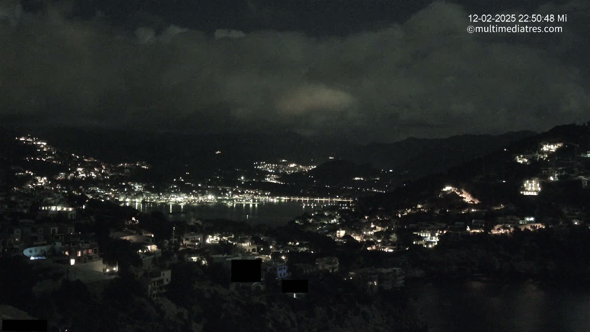 A nighttime view of a coastal town nestled on hillsides, illuminated by scattered lights, under a cloudy sky.
