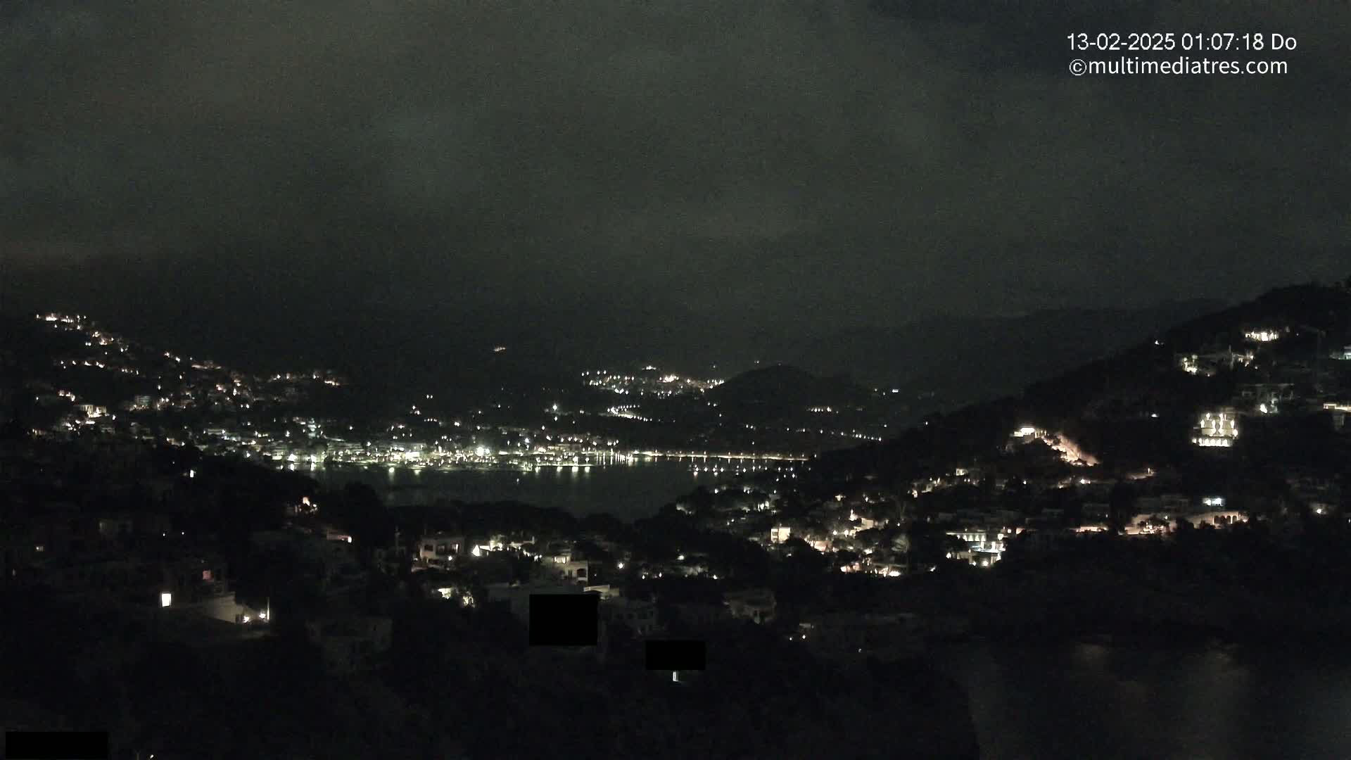 A nighttime view of a coastal town nestled on hillsides, illuminated by scattered lights, under a dark, cloudy sky.