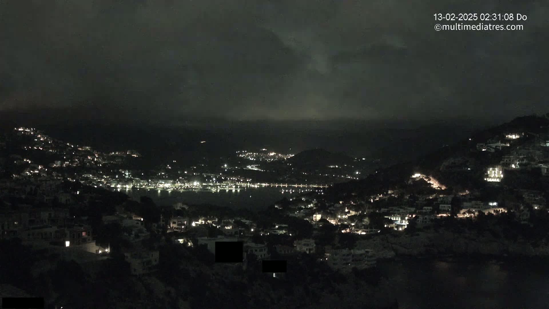 A nighttime view of a coastal town with scattered lights under a dark, cloudy sky.