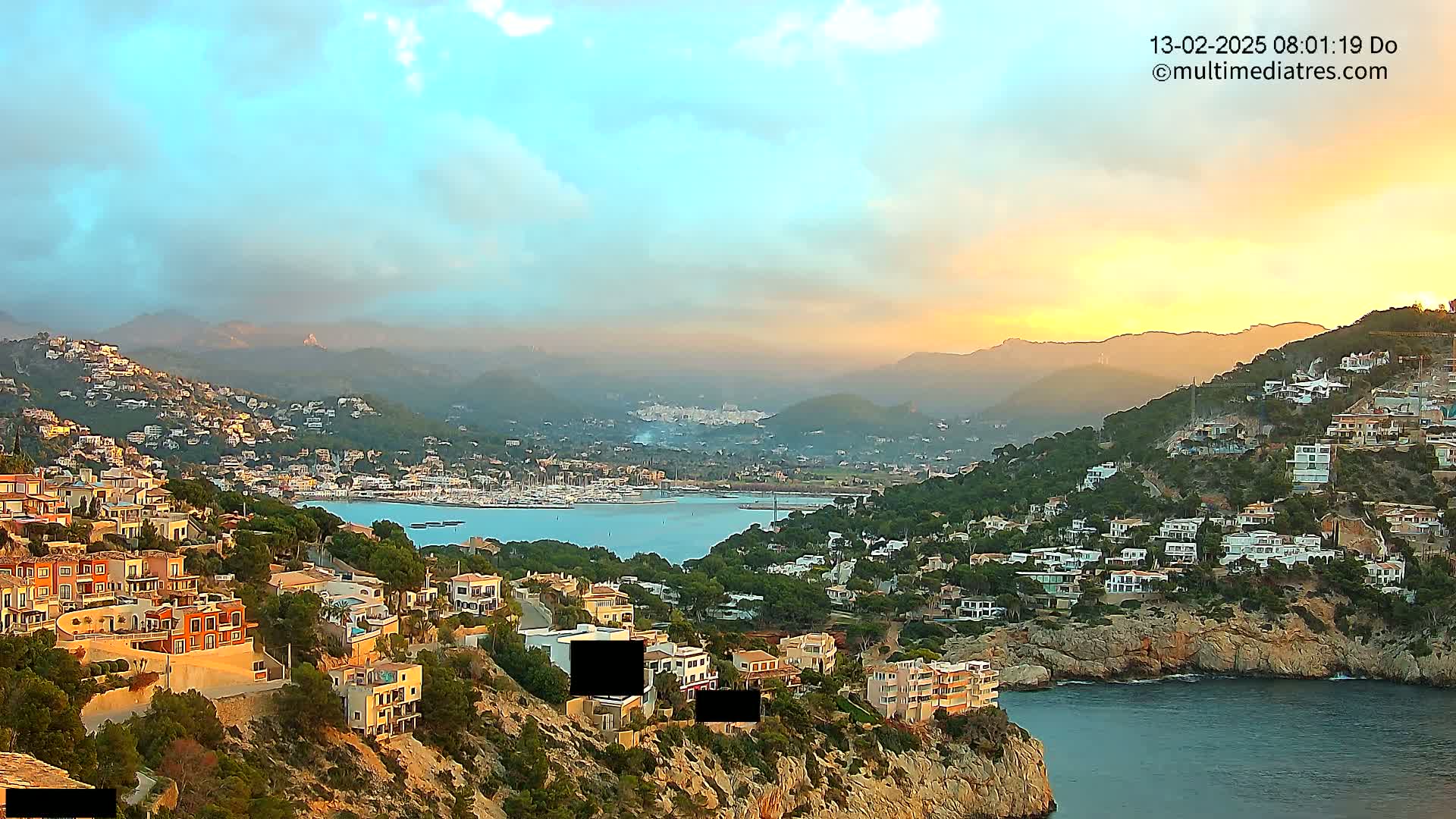A coastal town nestled on a hillside overlooking a calm bay at sunrise, with hazy mountains in the background.