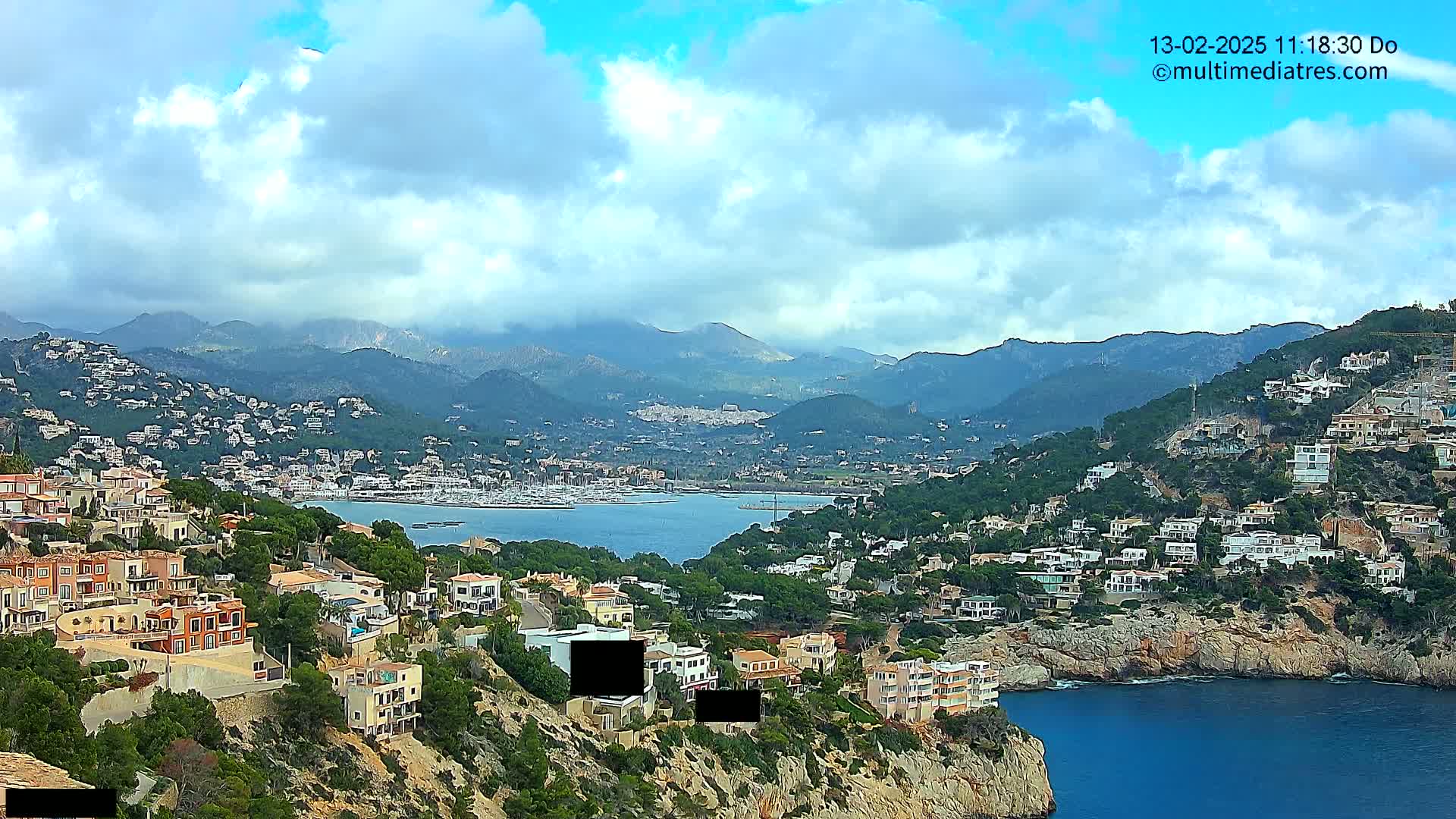 A coastal town nestled on a hillside overlooking a calm bay, with mountains in the background under a partly cloudy sky.