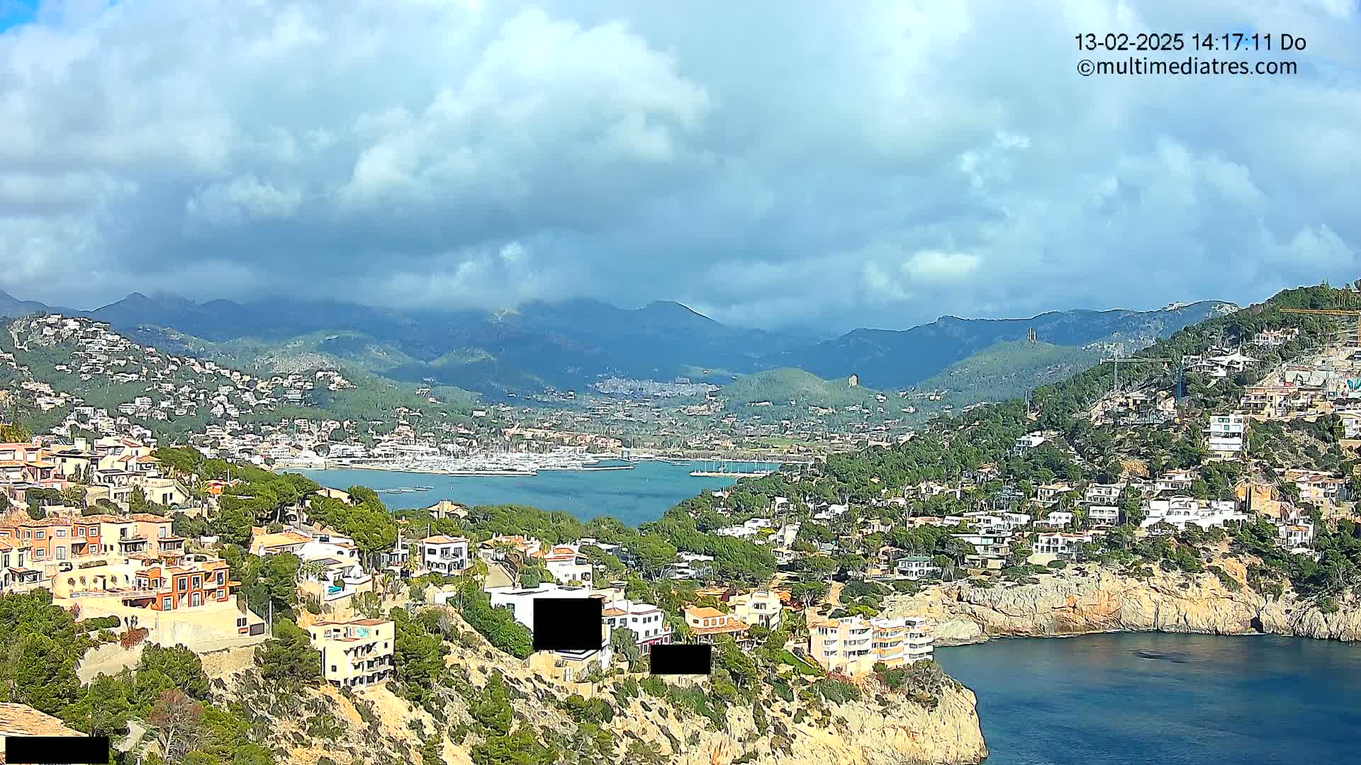 A coastal town nestled on a hillside overlooking a calm bay with a mountain range in the background under a partly cloudy sky.