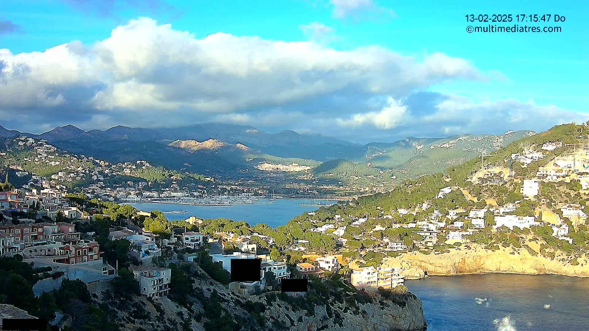 A coastal town nestled between mountains under a partly cloudy sky overlooks a calm bay filled with boats.