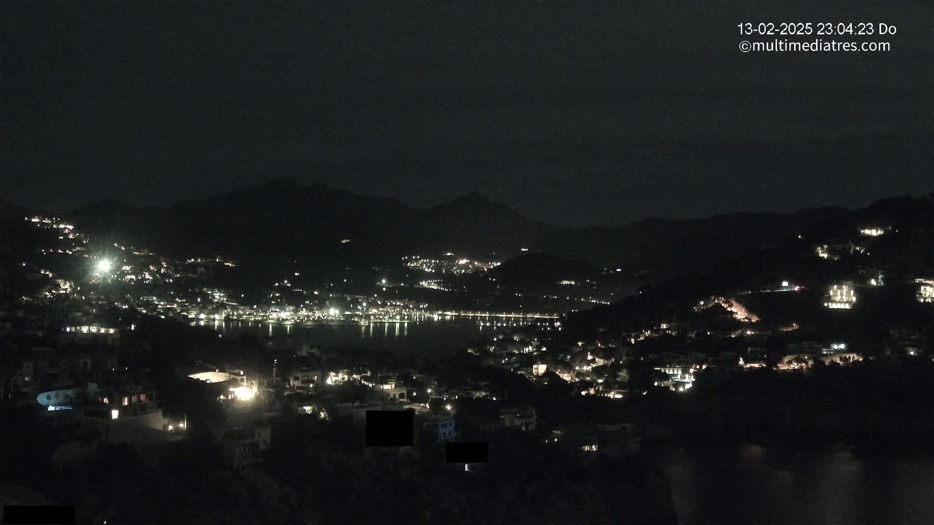 A nighttime view shows a coastal town nestled between dark hills, its lights reflecting on a calm body of water under a clear sky.