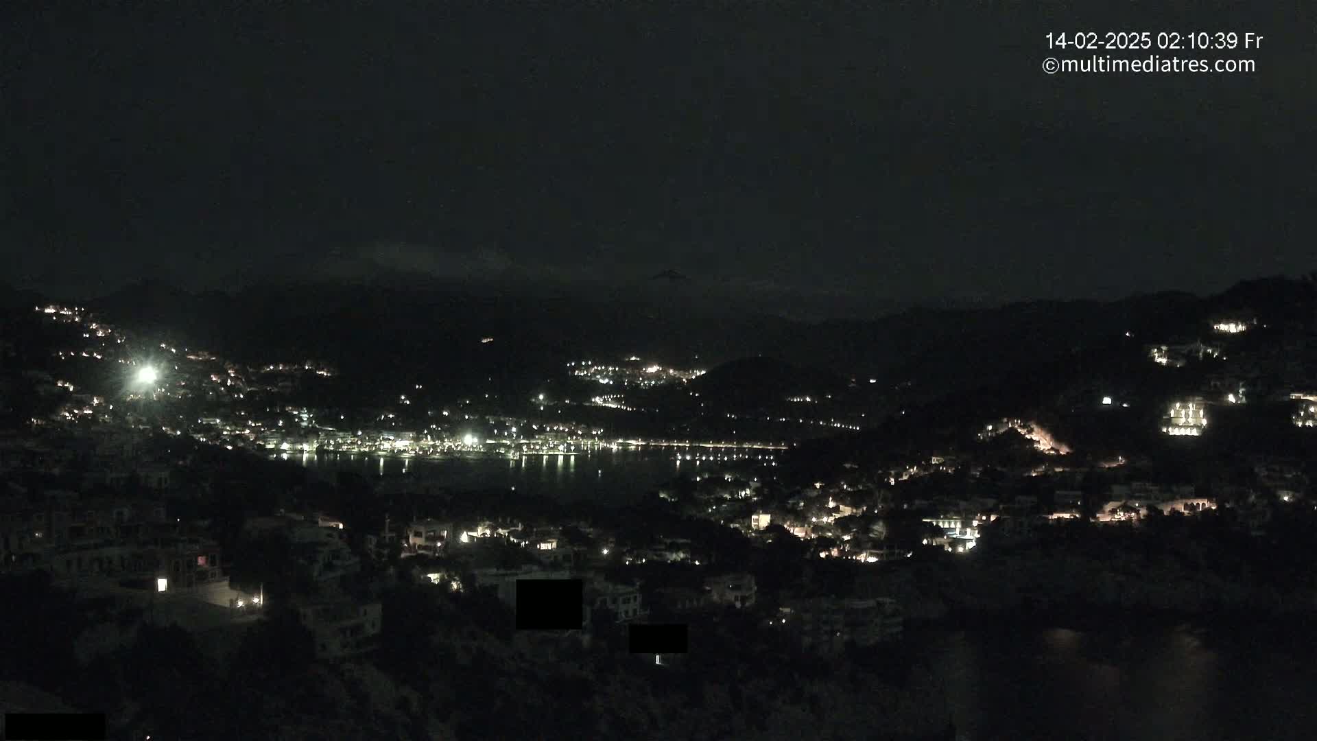 A nighttime view of a coastal town nestled between hills, with scattered lights illuminating the buildings and a dark body of water visible in the distance.