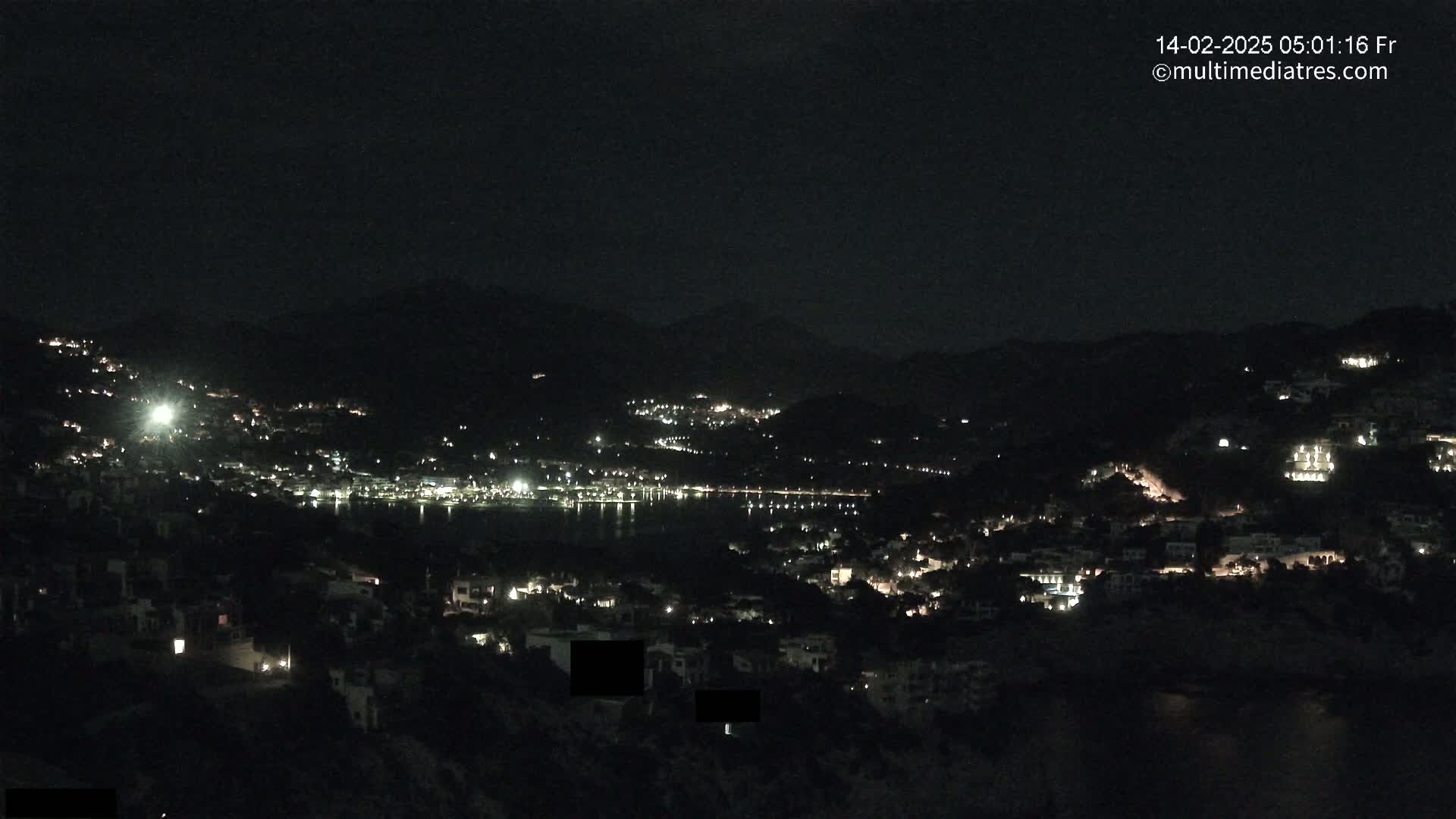 A nighttime view of a coastal town nestled between dark hills, with scattered lights illuminating buildings and streets along a dark body of water.