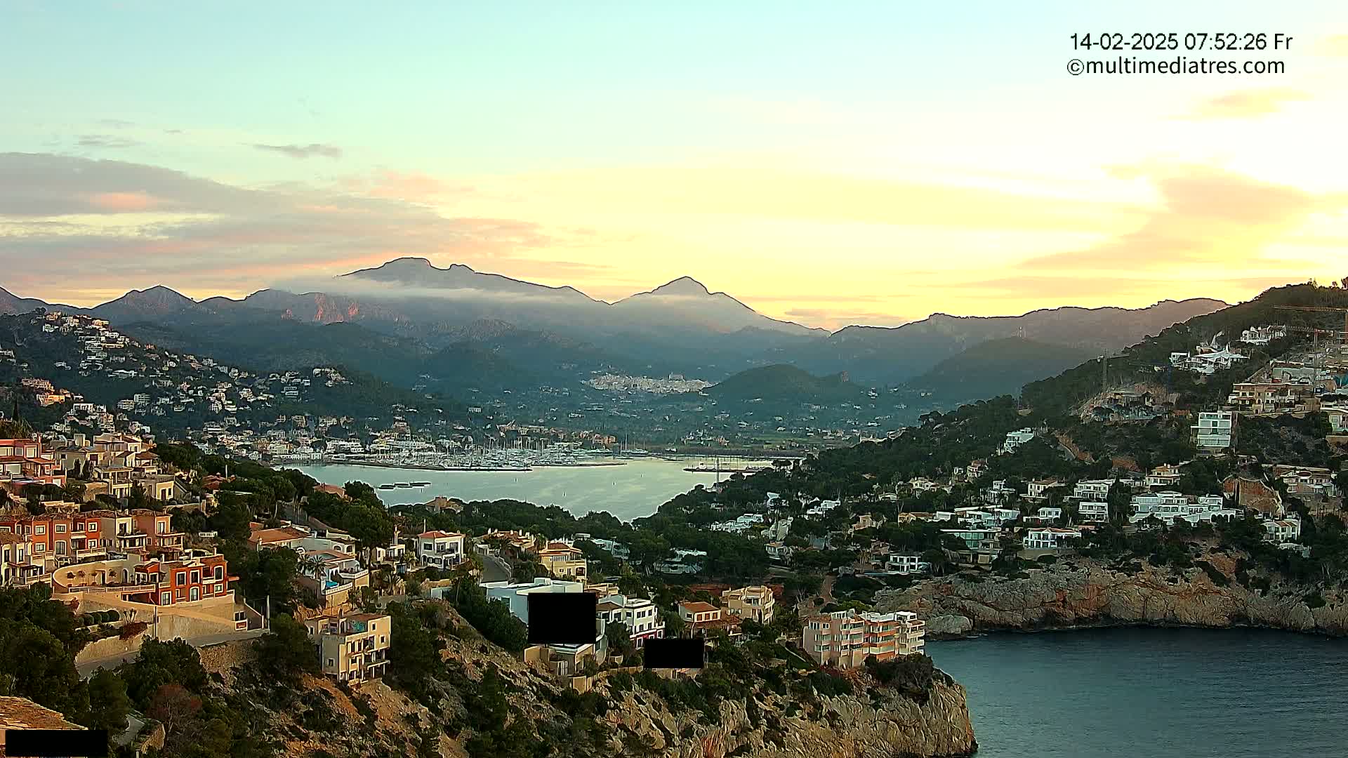 A coastal town nestled on a hillside overlooking a calm bay, with mountains in the background under a sunrise sky.
