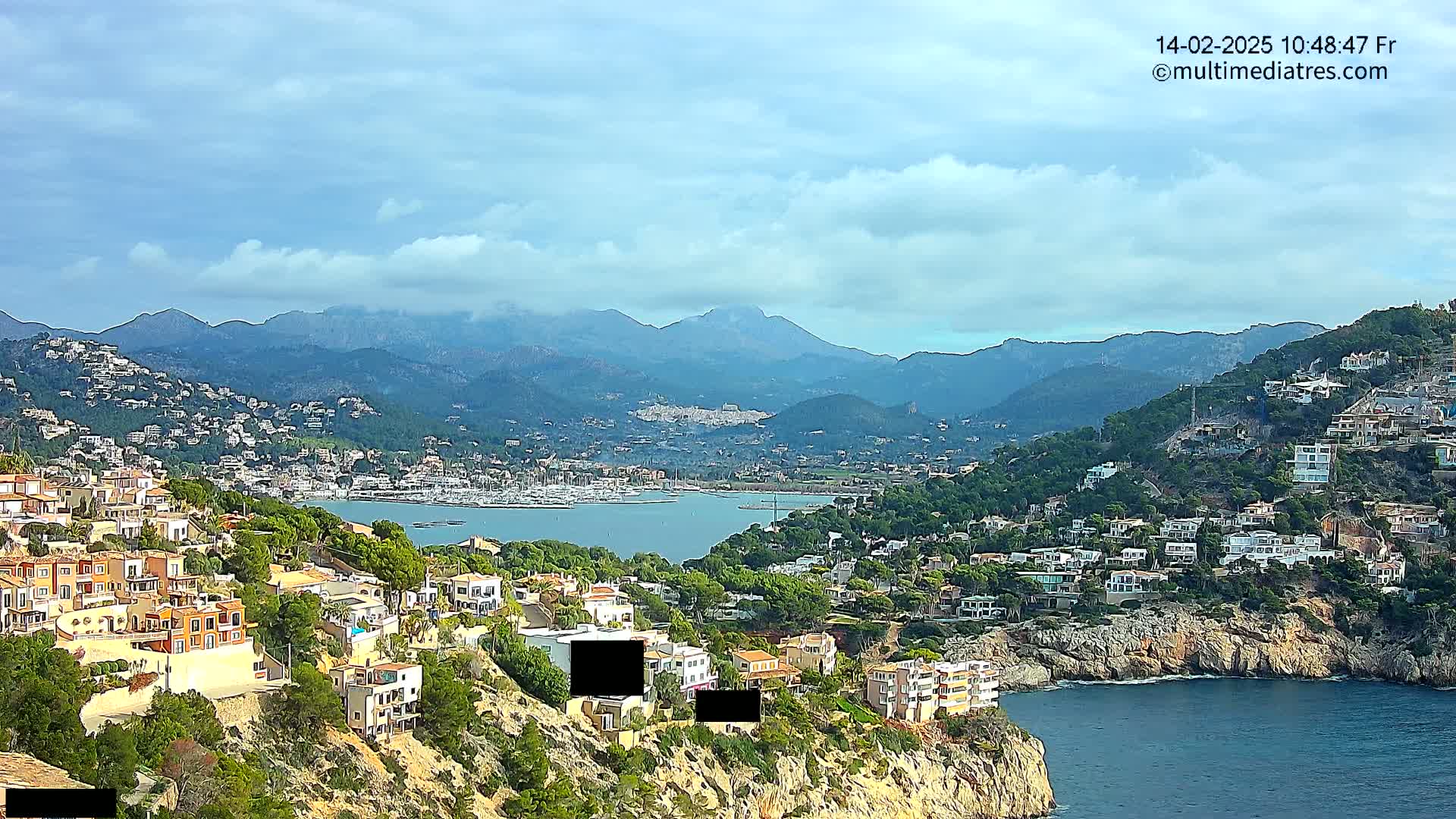 A coastal town nestled on a hillside overlooking a calm bay, with mountains in the background under a partly cloudy sky.