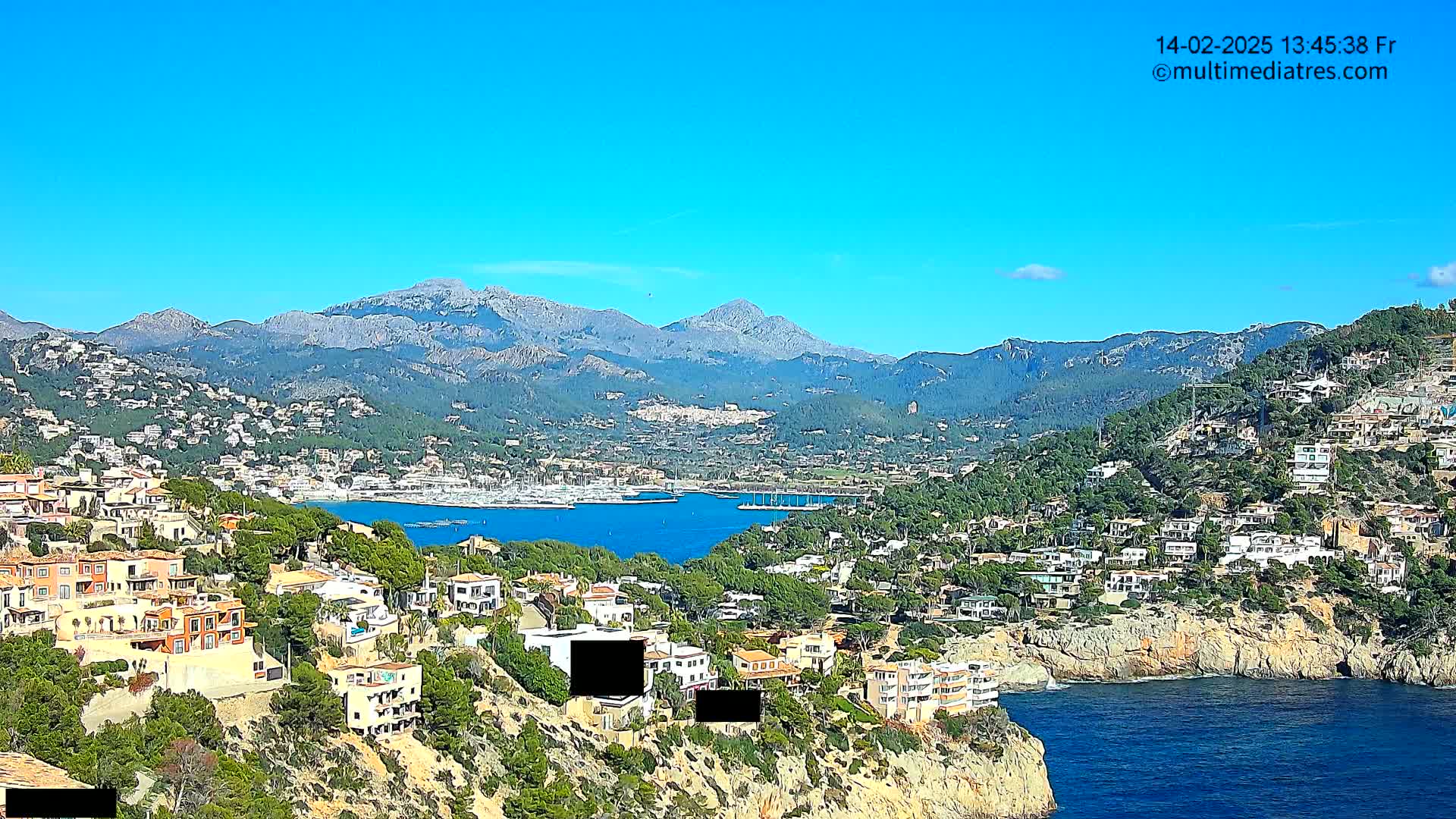 A coastal town nestled in a mountainous region, with a clear blue sky and calm waters in a harbor filled with boats.