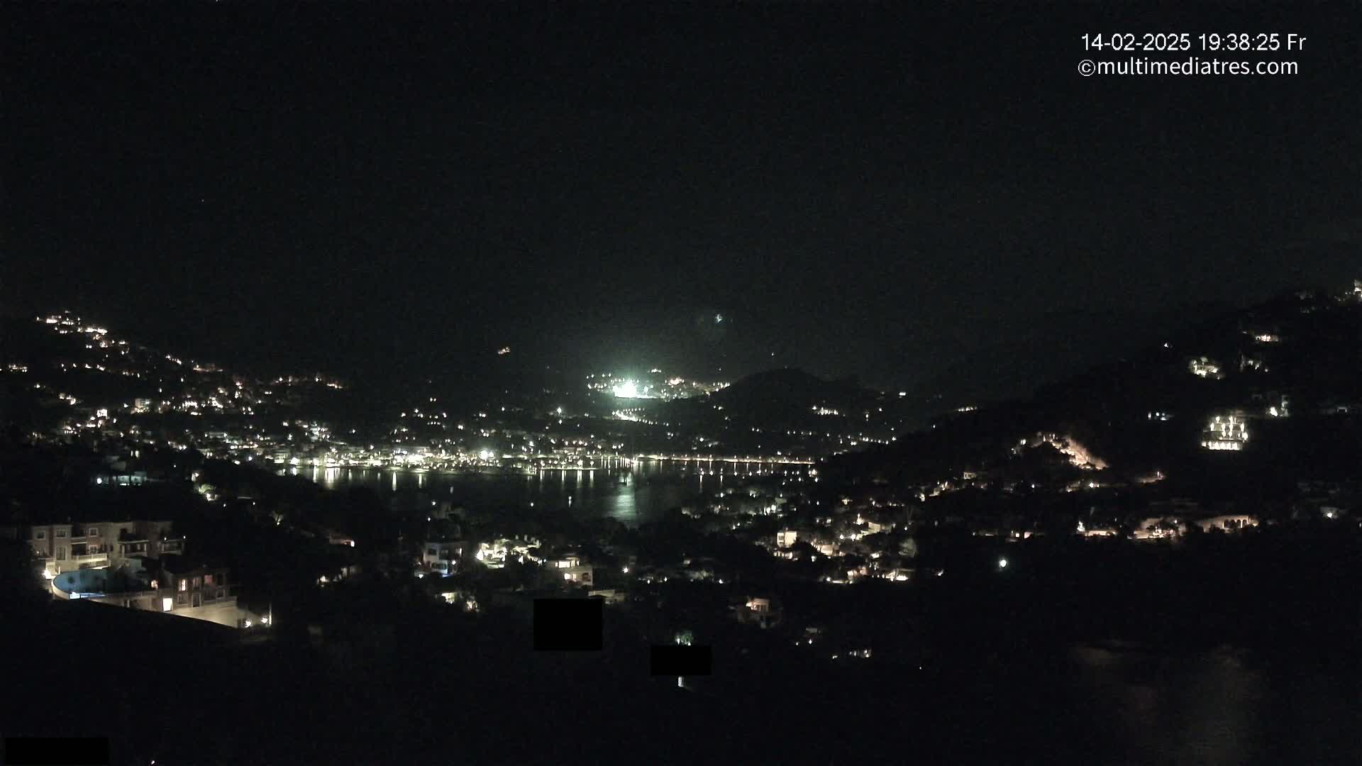 A nighttime view of a coastal town illuminated by scattered lights, nestled between hills and overlooking a calm body of water.
