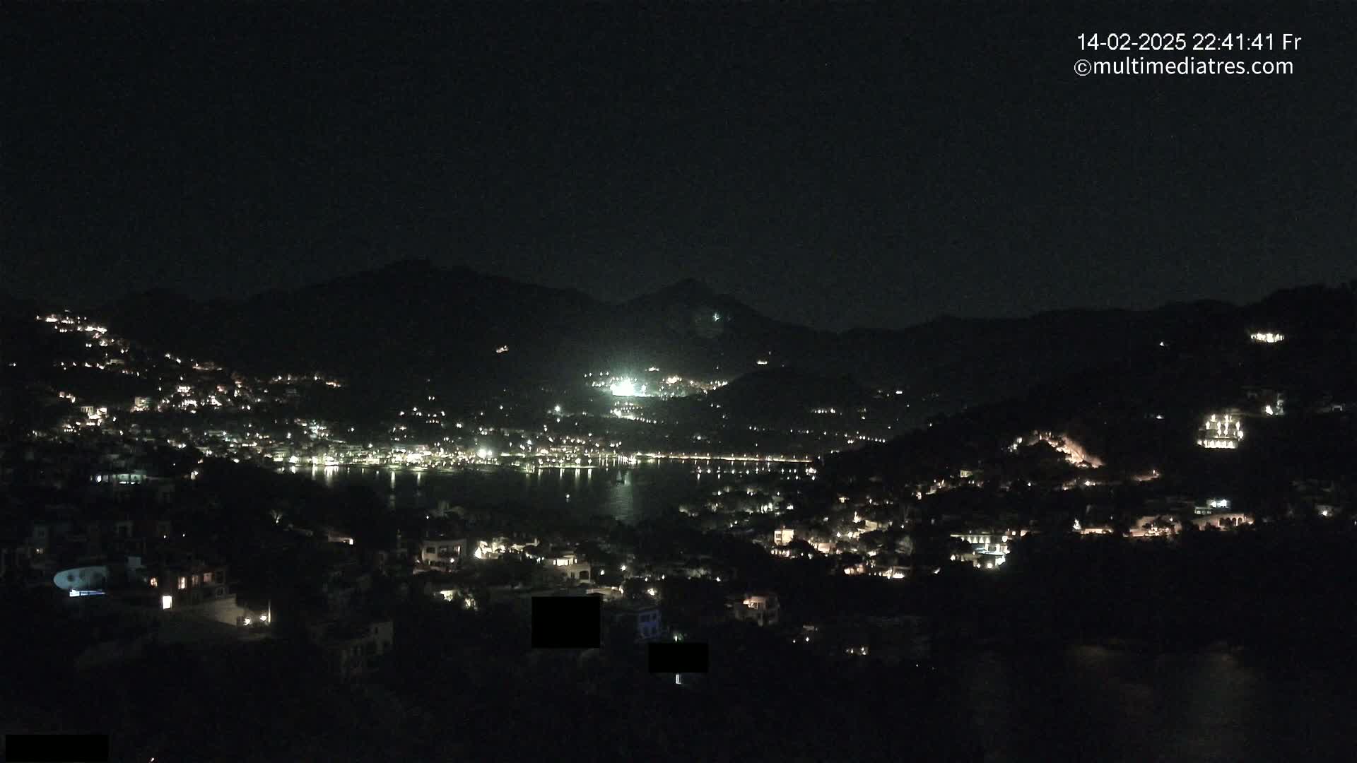 A nighttime view of a town nestled between hills and a body of water, illuminated by scattered lights.