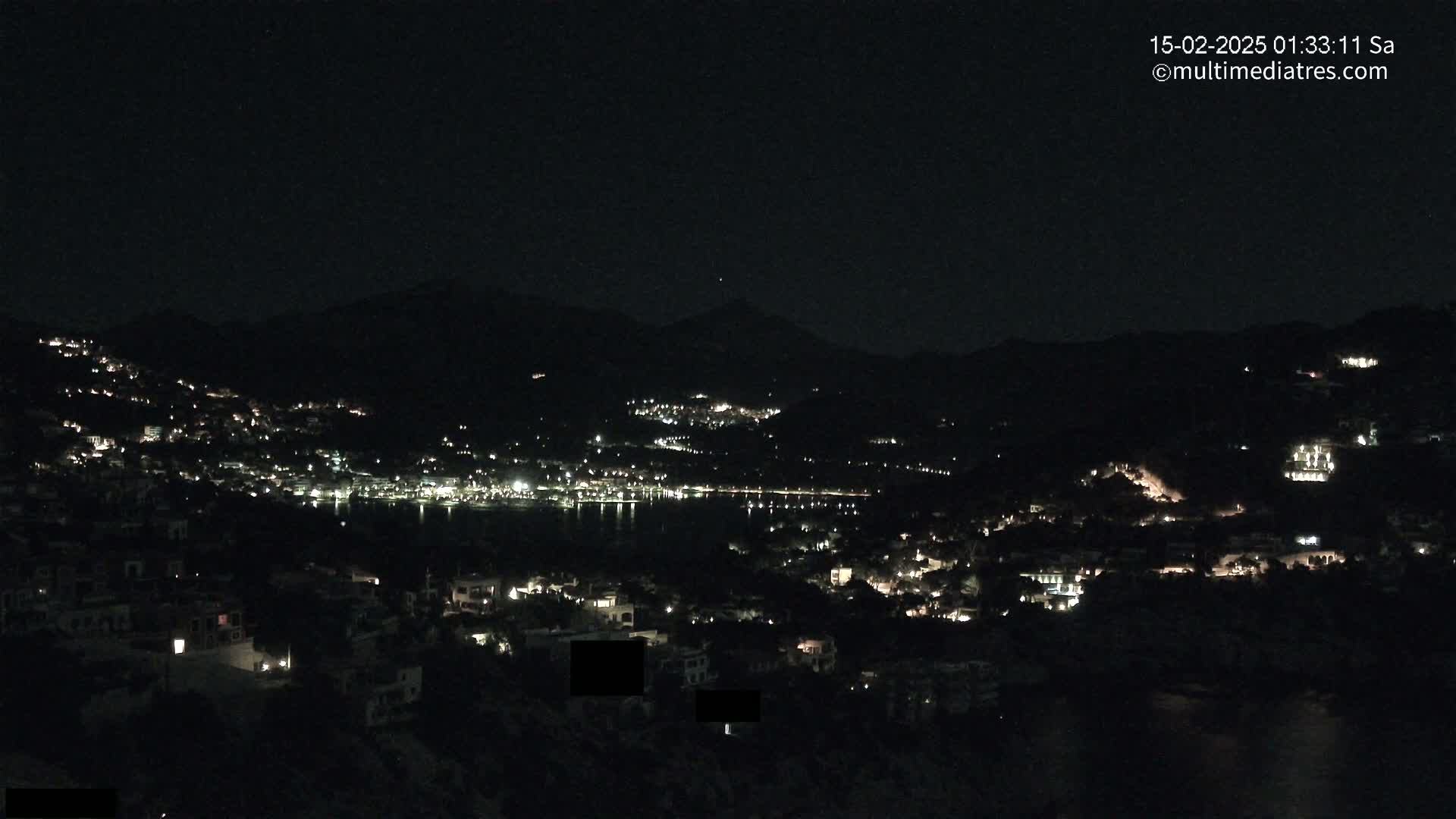 A nighttime view of a coastal town illuminated by scattered lights, nestled between dark hills and a calm body of water under a clear, dark sky.