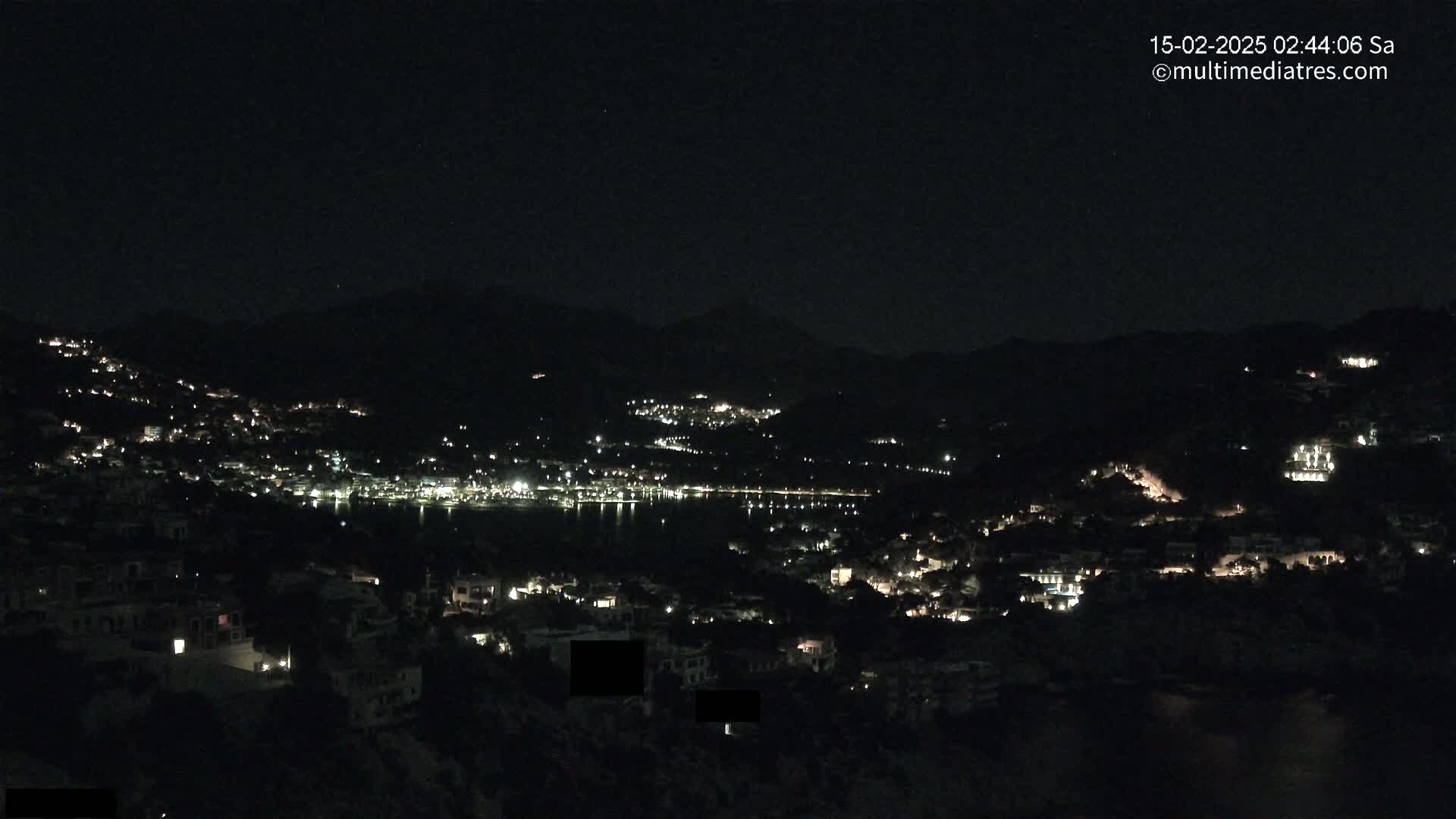 A nighttime view shows a coastal town illuminated with scattered lights, nestled between dark hills under a clear, starless sky.