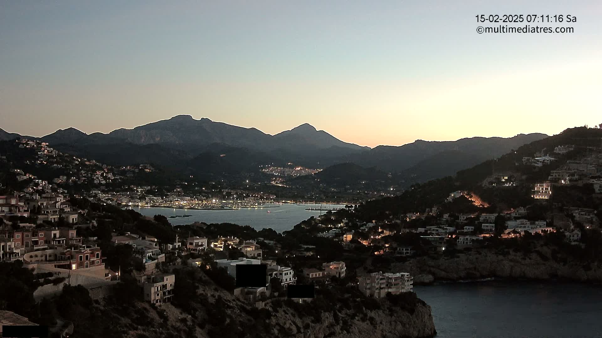 A coastal town nestled between hills and mountains at twilight, with the lights of buildings illuminated against a calm body of water.