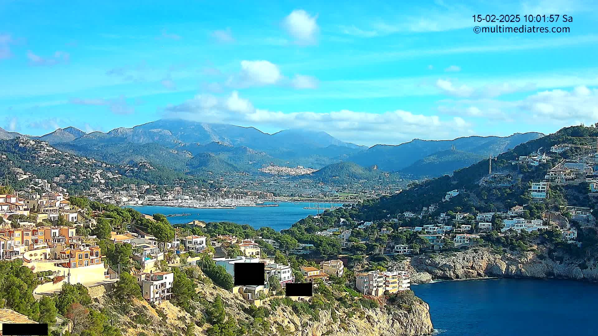 A coastal town nestled on a hillside overlooks a calm bay filled with sailboats, with mountains in the background under a mostly sunny sky.